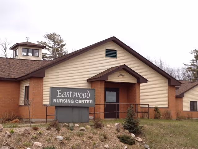 Exterior view of Eastwood Nursing Center Inc building with a beige and brown facade, a sign displaying the facility name, and some landscaping with rocks and small bushes in front.