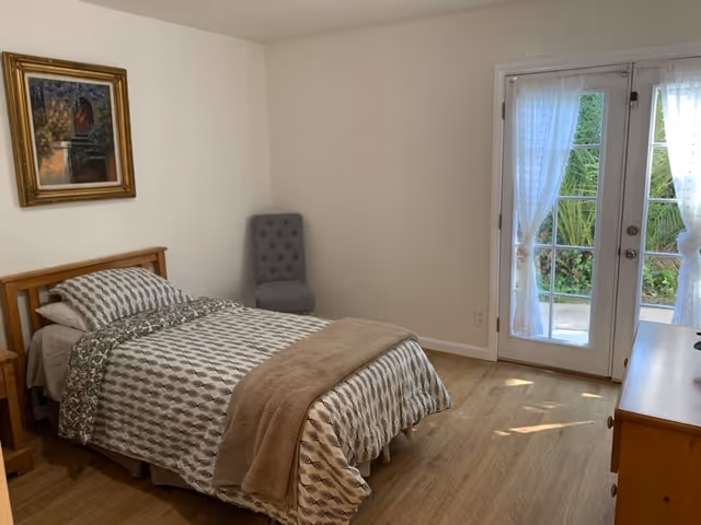 A simple bedroom with a single bed covered in patterned bedding and a beige blanket. There is a gray upholstered chair in the corner, a framed painting on the wall, and a wooden dresser. French doors with sheer white curtains open to a garden outside, allowing natural light to fill the room.