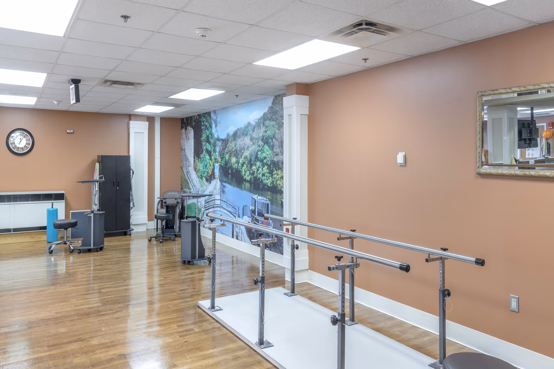 A rehabilitation or physical therapy room with parallel bars for walking exercises, chairs, a clock on the wall, and a large mural depicting a scenic river and greenery. The room has wooden flooring and peach-colored walls with a mirror on one side.