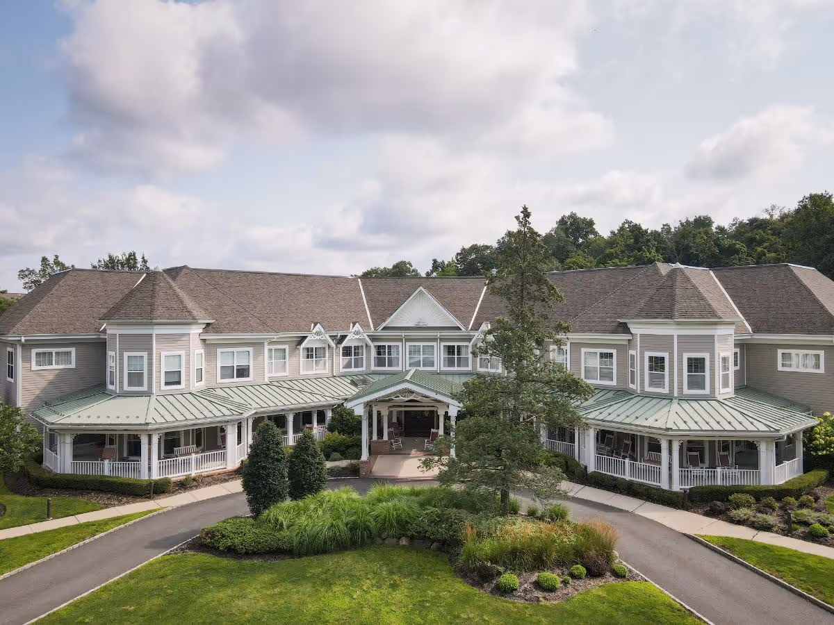Front exterior of a two-story assisted living building with wraparound porches, a green metal roof, and landscaped driveway.