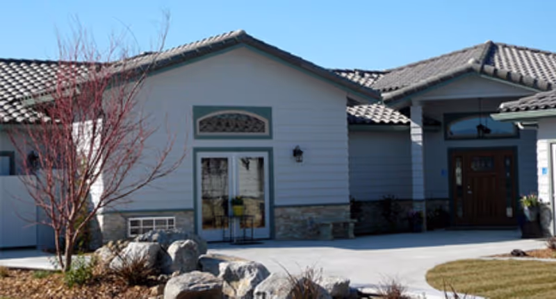 Exterior view of a single-story building with a tiled roof, light-colored siding, and stone accents near the base. There is a small tree with no leaves and some landscaping rocks in the front yard. The entrance features a wooden door with glass panels and a covered porch area.