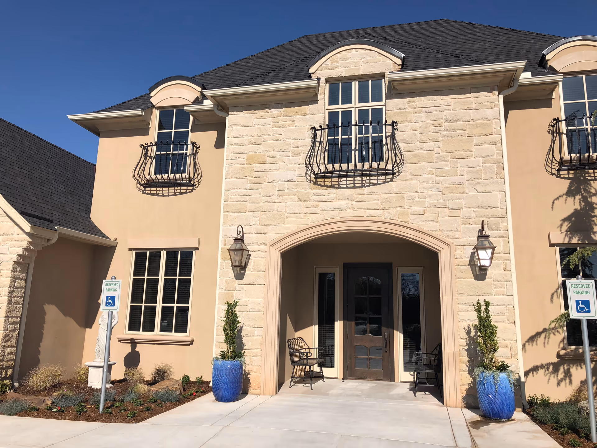 Beige stone senior living facility entrance with arched doorway, chairs, planters, and reserved parking.