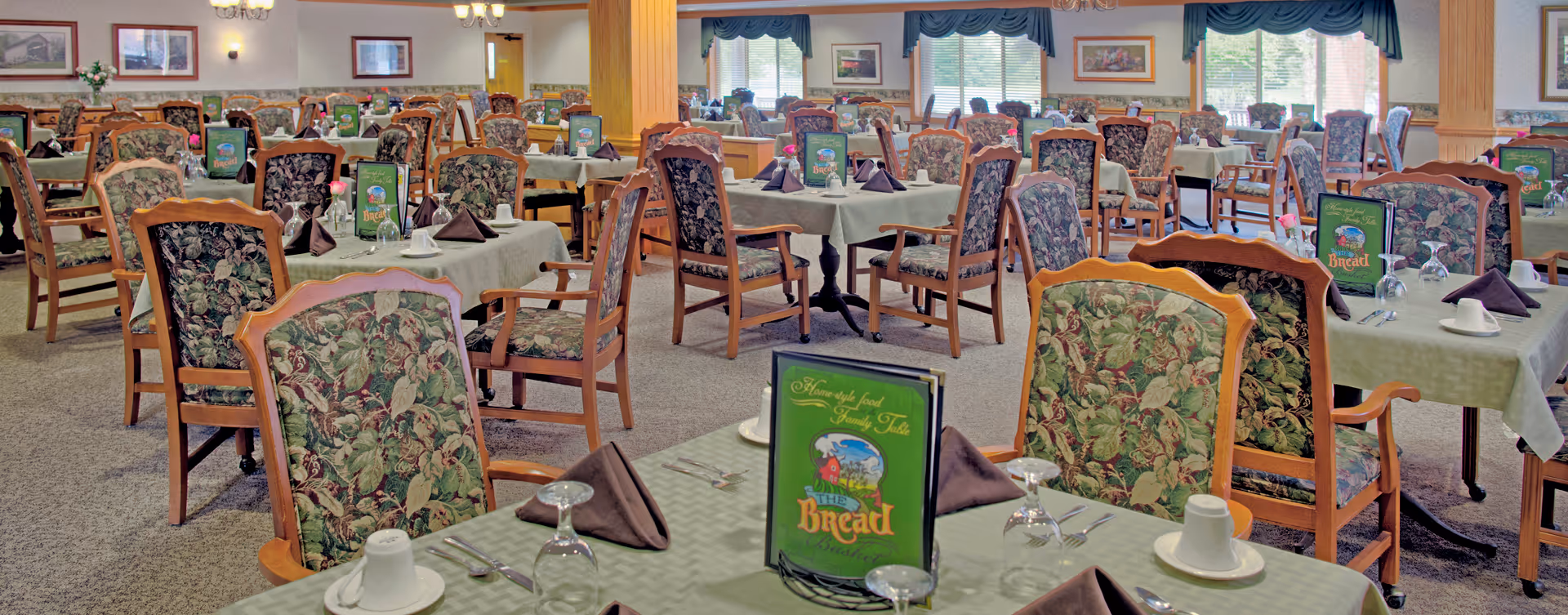 A spacious dining room with multiple tables covered in light green tablecloths, each set with brown folded napkins, white cups, glasses, and silverware. The chairs have wooden frames with floral patterned upholstery. Menus titled 'Bread' are placed on each table. The room has large windows with blue valances, framed pictures on the walls, and carpeted flooring.