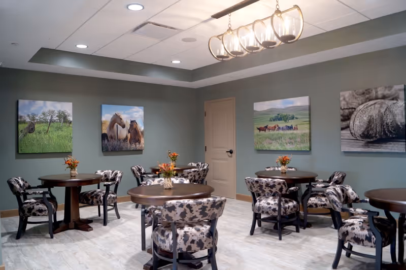 Small dining room with round wooden tables, patterned upholstered chairs, floral centerpieces, and landscape artwork on muted green walls under a chandelier.