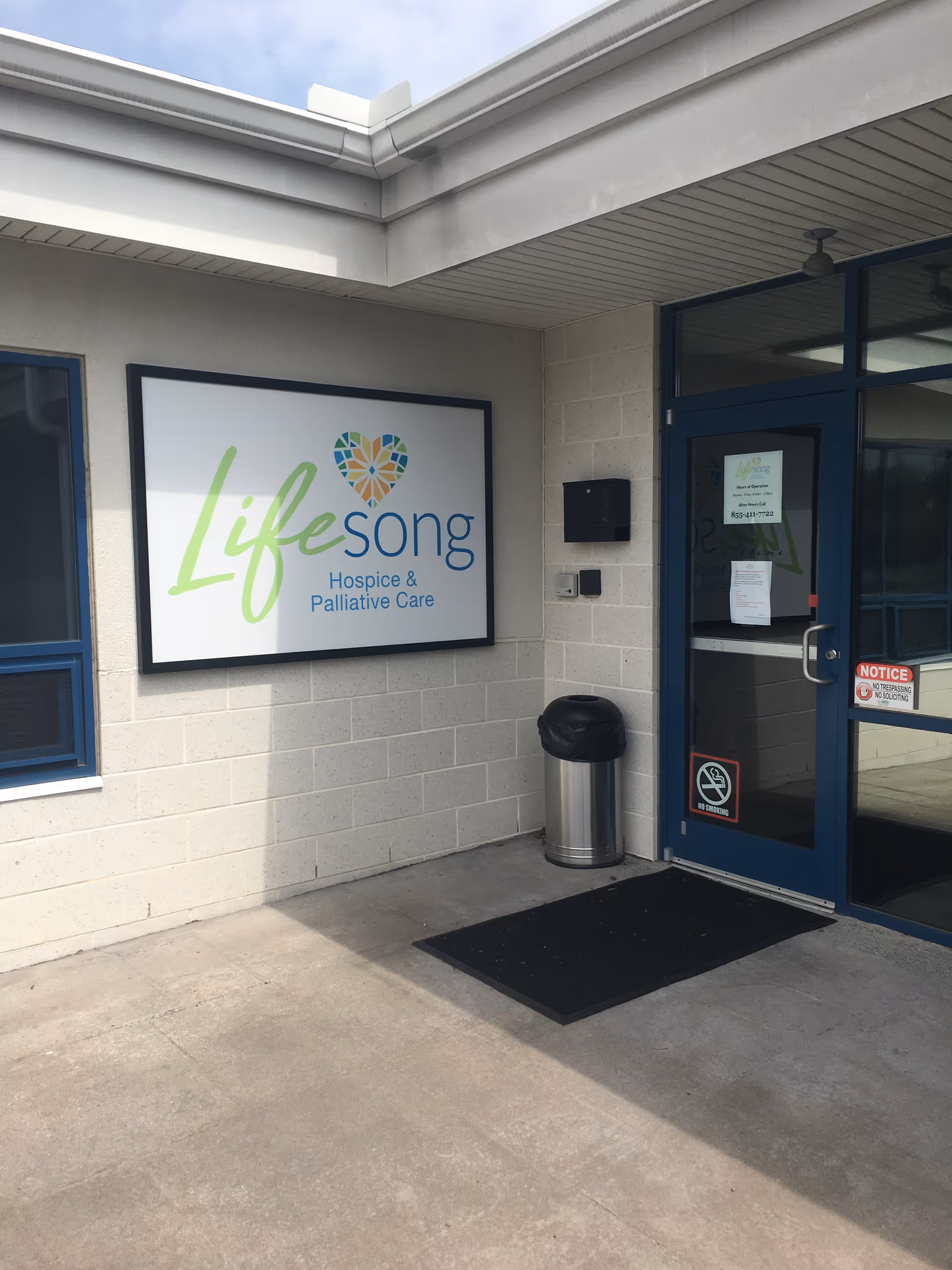 Exterior entrance of Lifesong Hospice & Palliative Care with sign, doorway, and trash can.