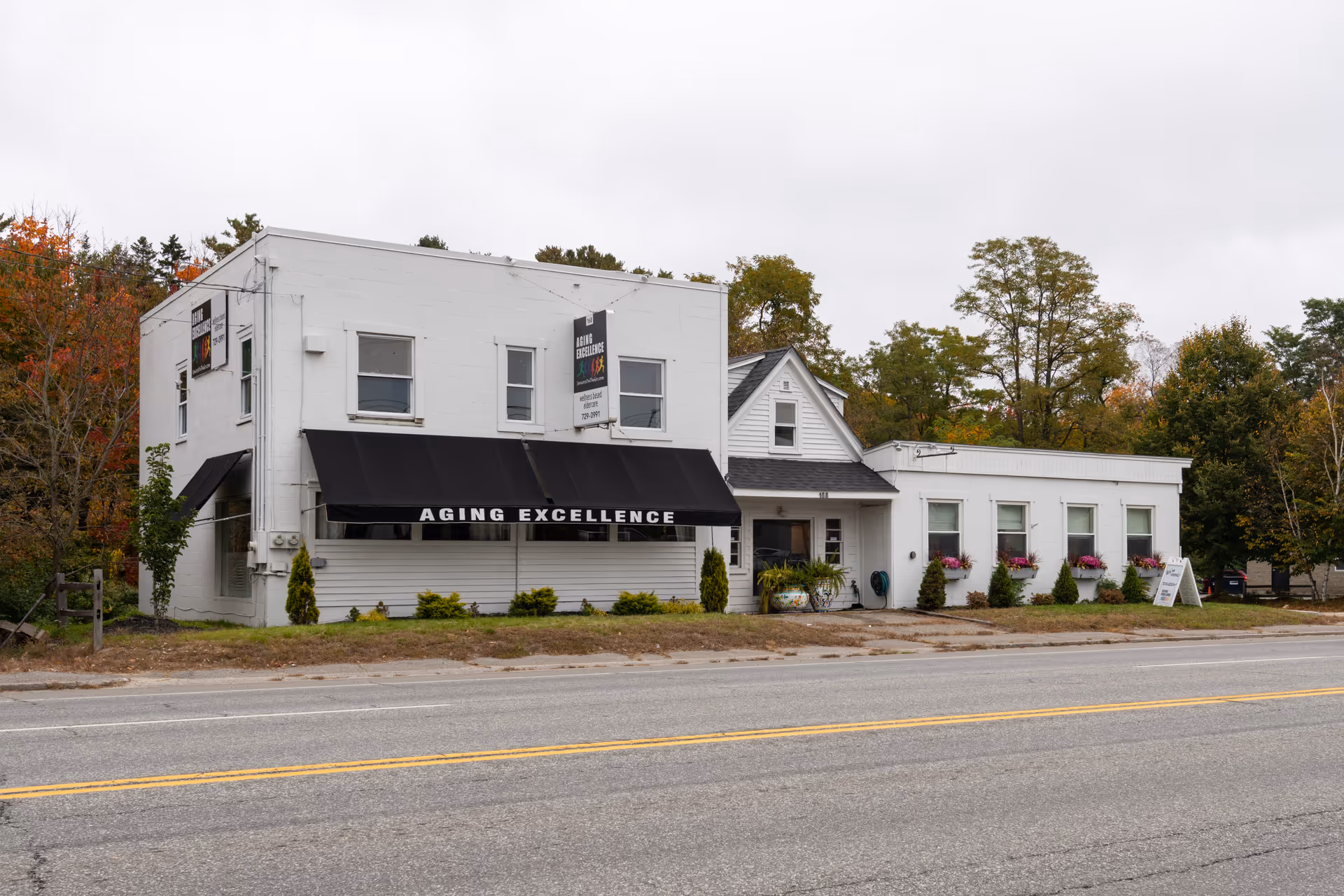 White two-story senior facility with a black awning and 'Aging Excellence' sign along the road