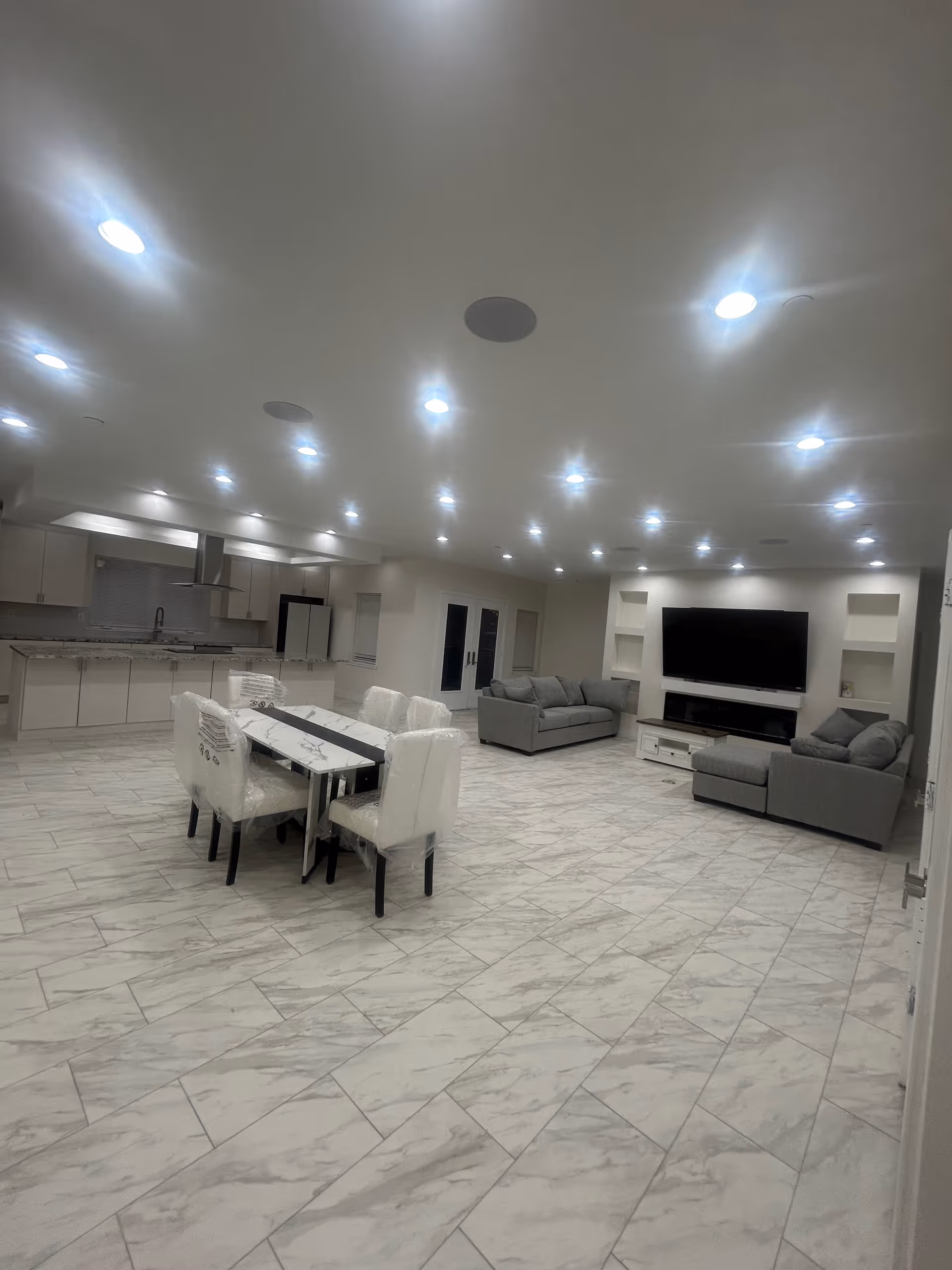 Open, modern living area with a dining table, white chairs, and gray sofas around a TV.