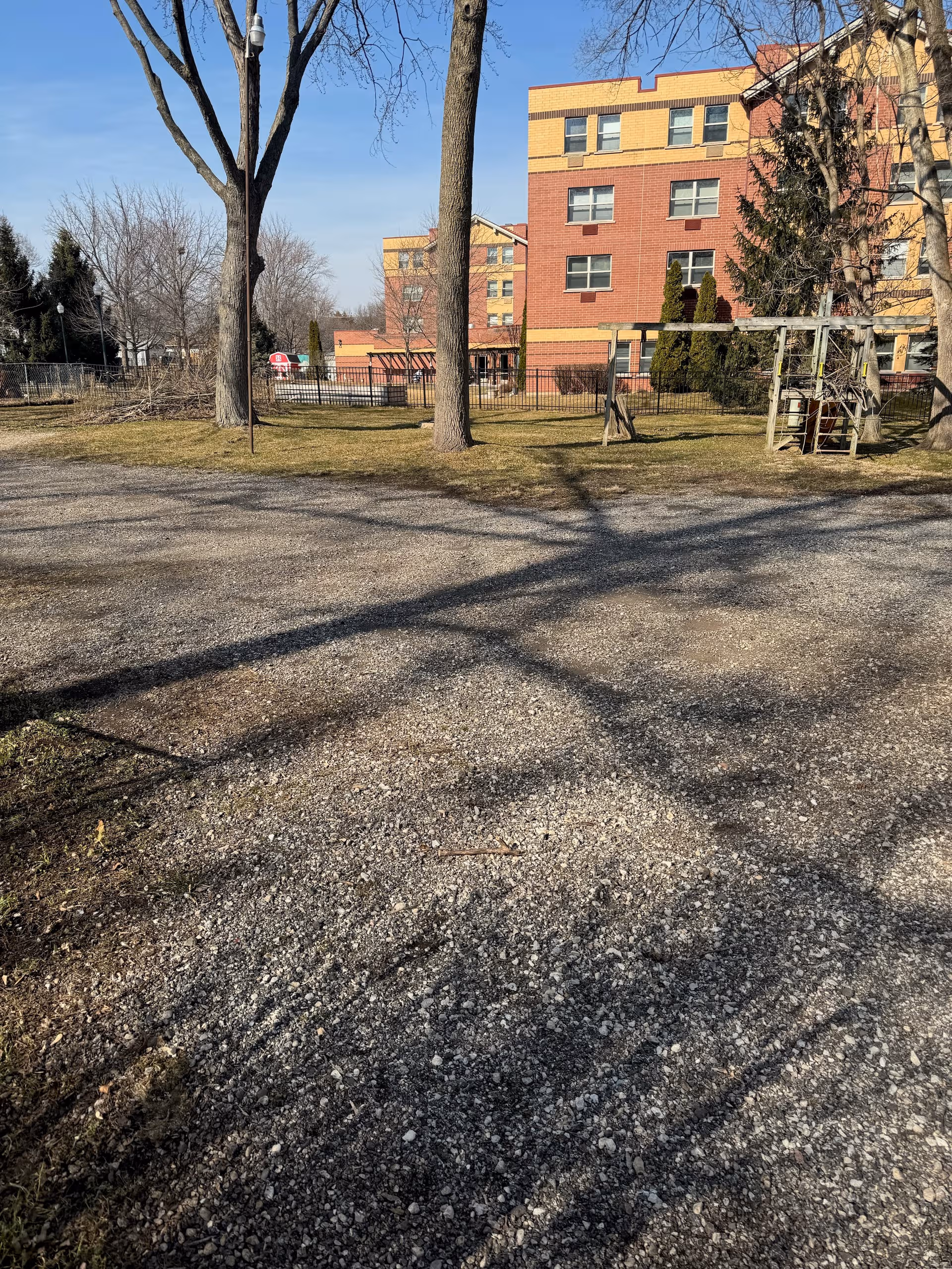 Outdoor area with trees, a playground, and a gravel path beside a brick building.