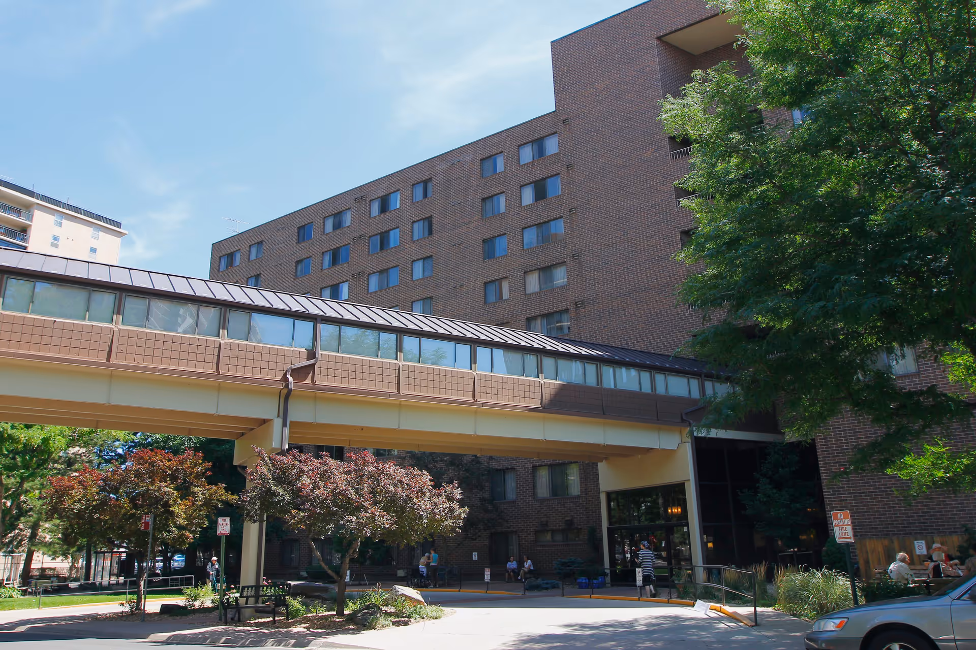 Front exterior of a multistory brick senior living building with a covered pedestrian skywalk over the driveway and trees in front.