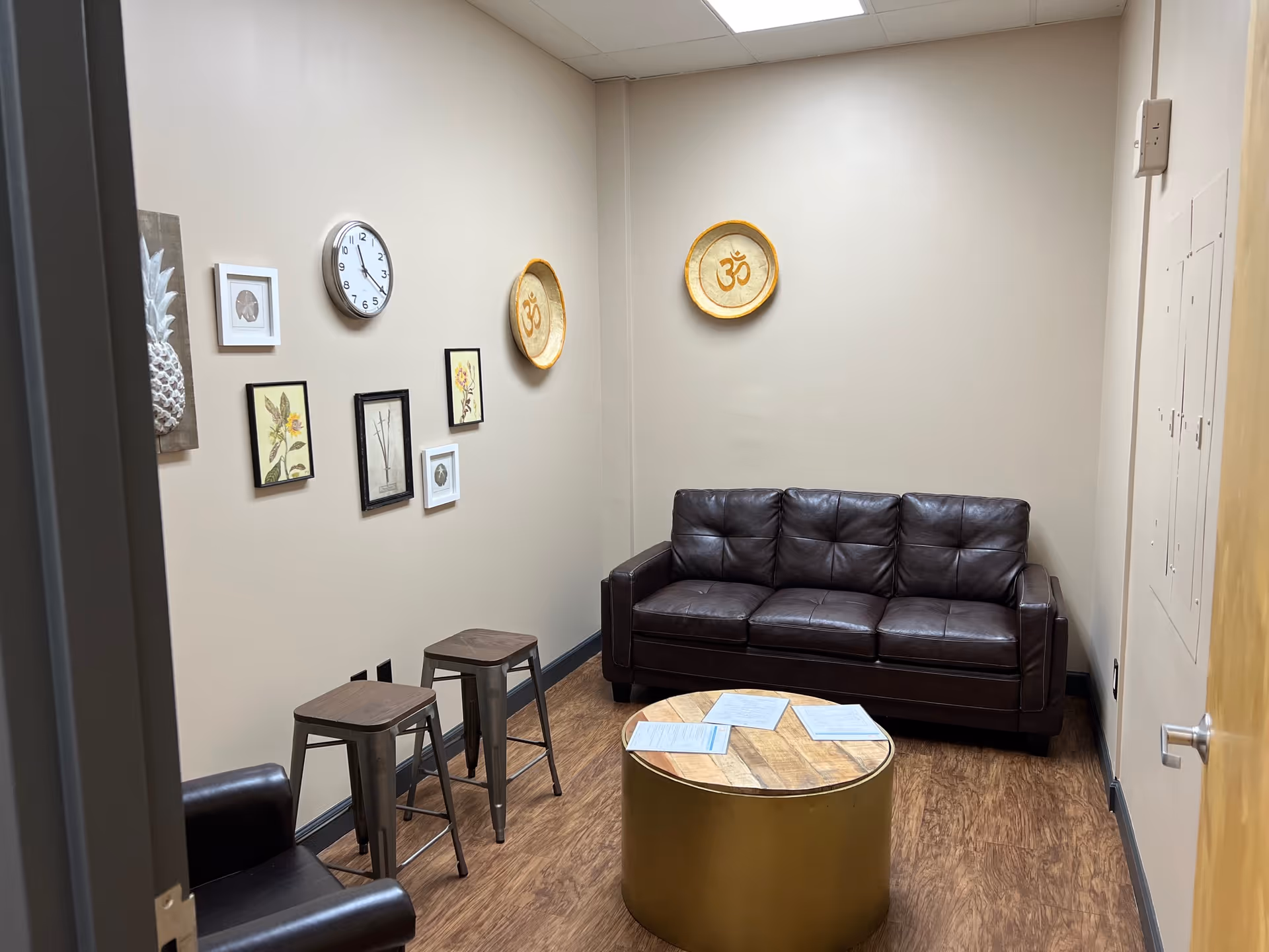 A small interior lounge with leather sofa, round coffee table, stools, wall decor, and a clock.