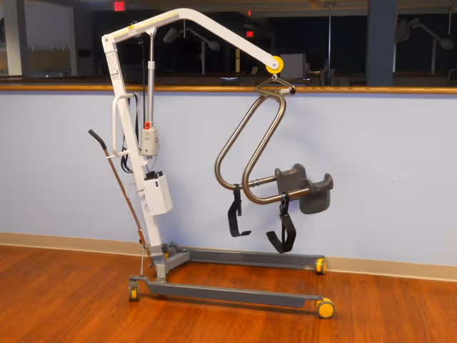 A floor-based patient lift stands in a resident care room with wooden floor.