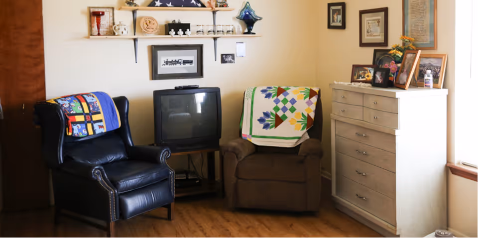 A cozy living room with two armchairs, a small TV, shelves, and a dresser.