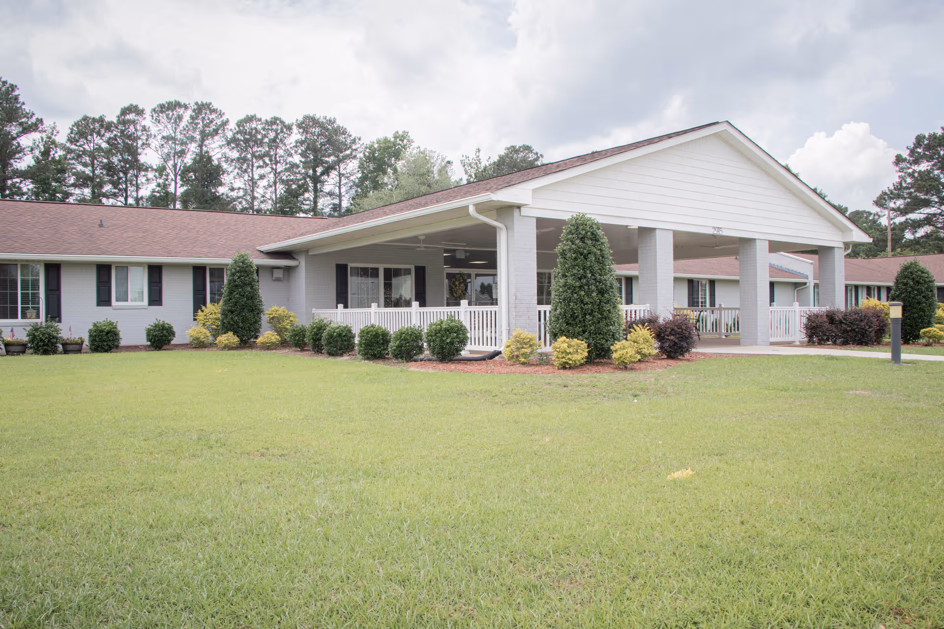 Exterior view of The Gardens of Trent senior living facility showing a single-story building with a covered entrance, white railings, and neatly trimmed bushes and trees in front. The building has a light gray facade with dark window shutters and a brown roof, set against a backdrop of tall pine trees and a partly cloudy sky.