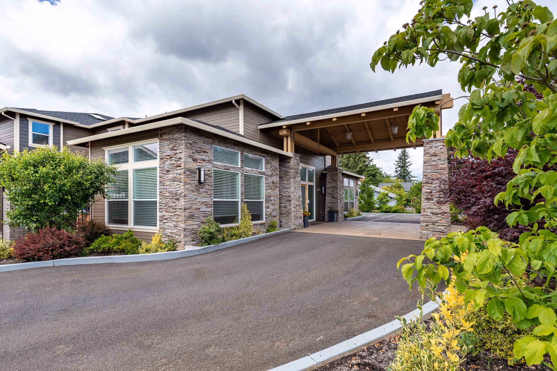 Entrance of a modern memory care building with a stone-clad facade, covered porte-cochère, driveway and landscaped shrubs.