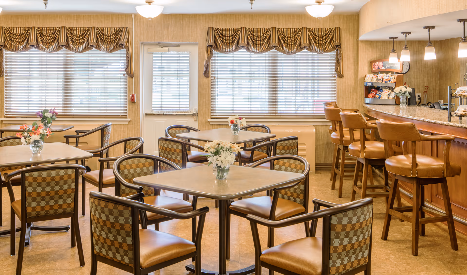 A bright dining room with square tables and patterned chairs, a small bar with stools, and windows with blinds and valances.