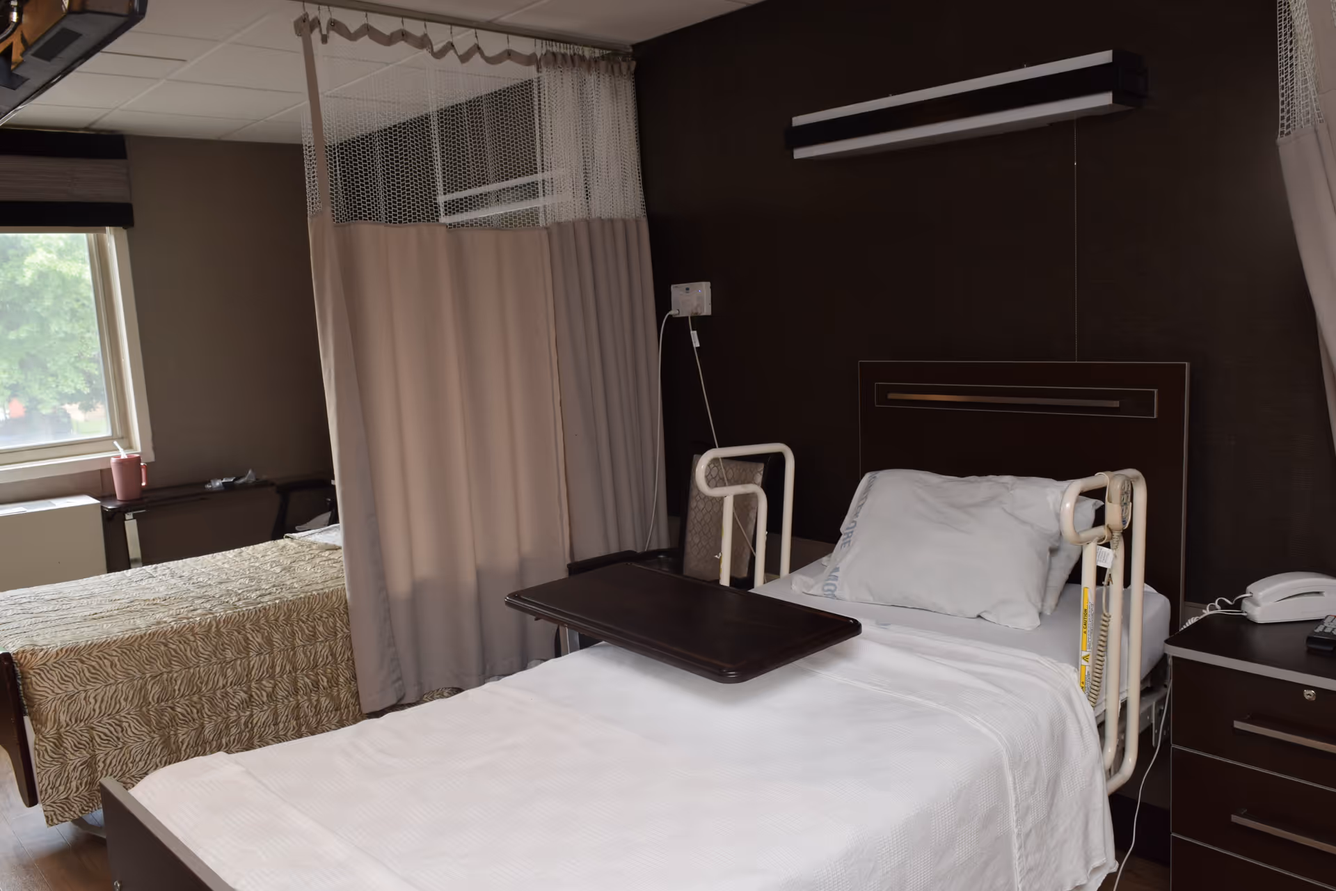 Hospital-style patient bedroom with two beds separated by a privacy curtain and bedside tables.