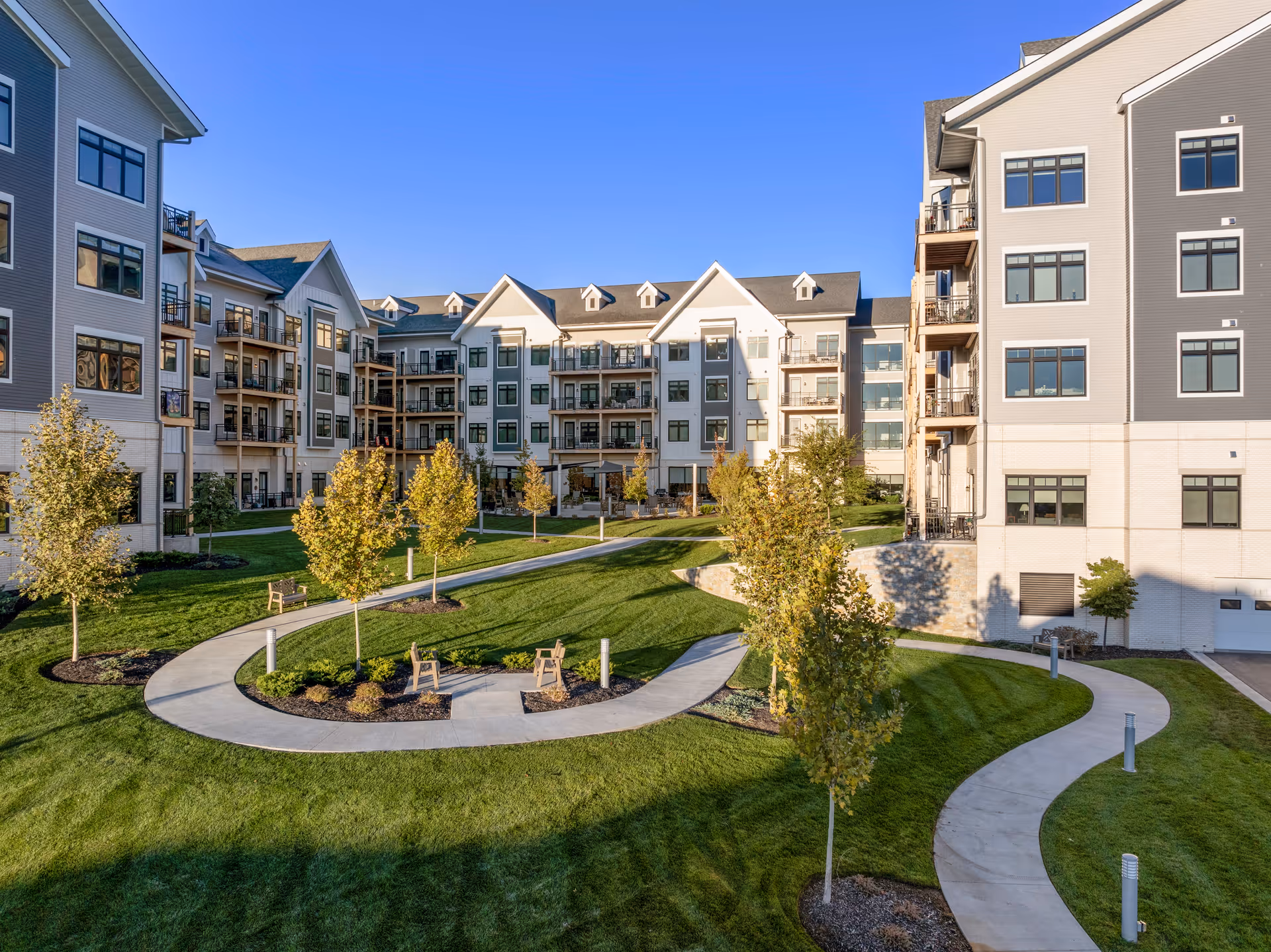 Outdoor courtyard area of a senior living facility with well-maintained green lawns, young trees, curved concrete walkways, benches, and multi-story residential buildings surrounding the space under a clear blue sky.