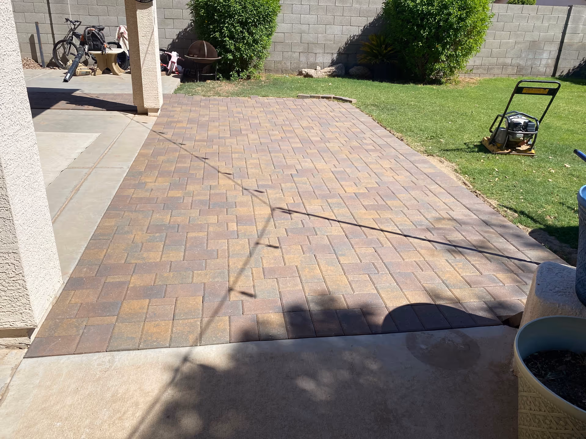 Paved brick patio beside a grassy yard with lawn equipment and bicycles.