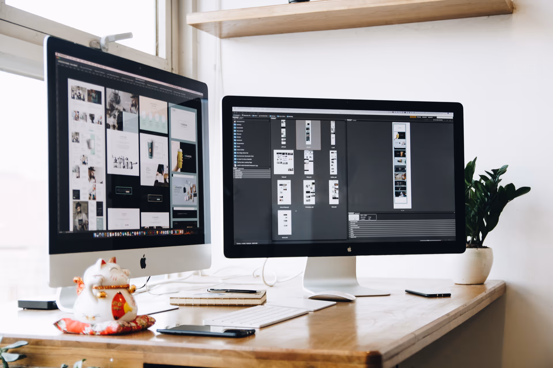 Two iMacs on a wooden desk with files on screens, plant, and decorative figurine.