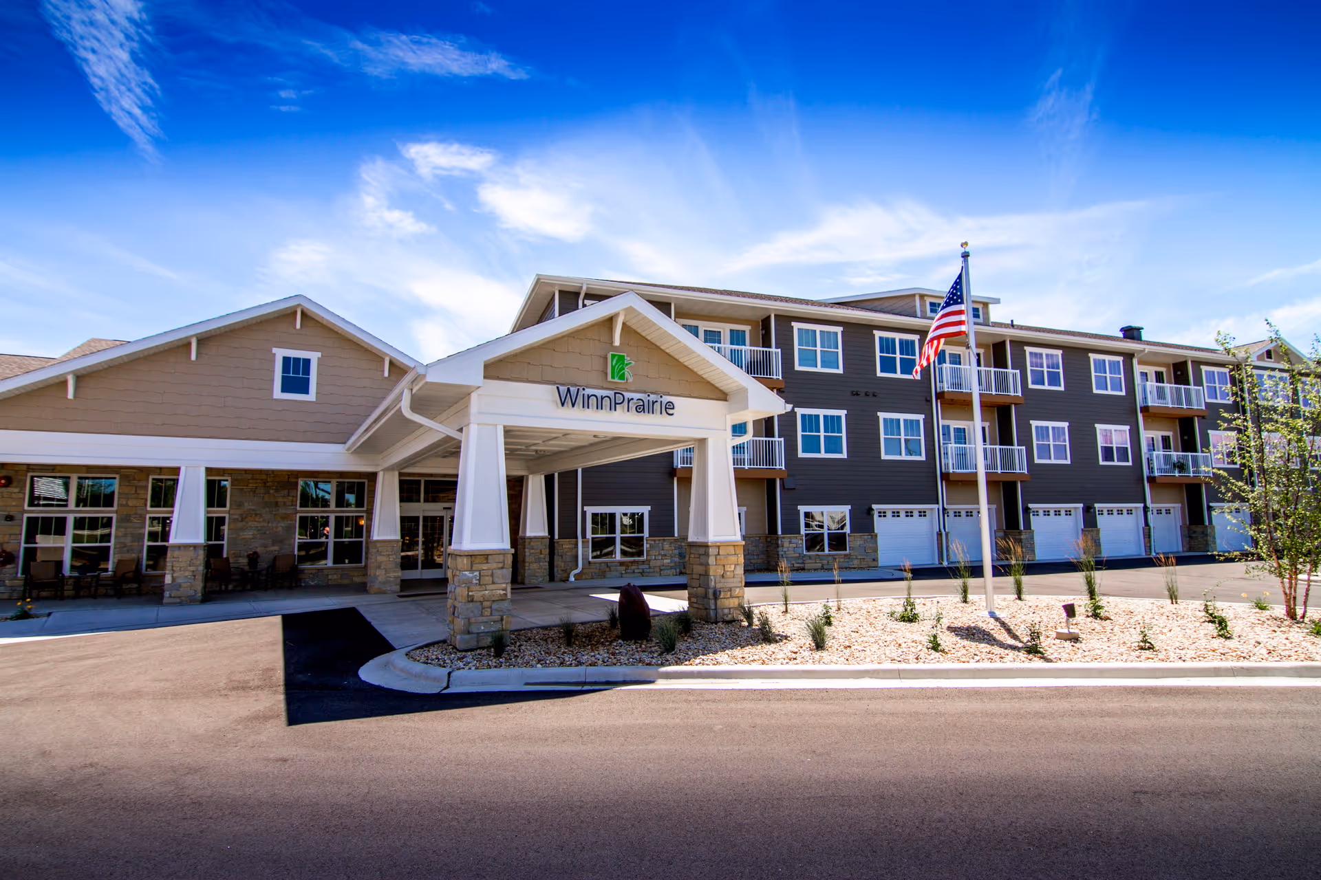 Exterior front of the Winn Prairie senior living building showing the covered entrance, American flag, and multi-story balconies under a blue sky.