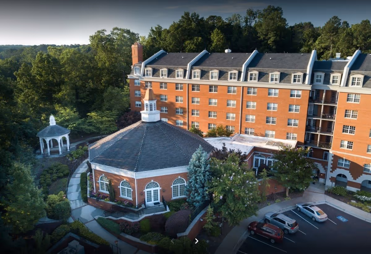 Aerial view of King's Bridge Retirement Community showing a large multi-story brick building with multiple windows and dormer windows on the roof. In front of the building is a smaller octagonal brick structure with a cupola on top. Surrounding the buildings are landscaped gardens, trees, a gazebo, and a parking area with several cars.