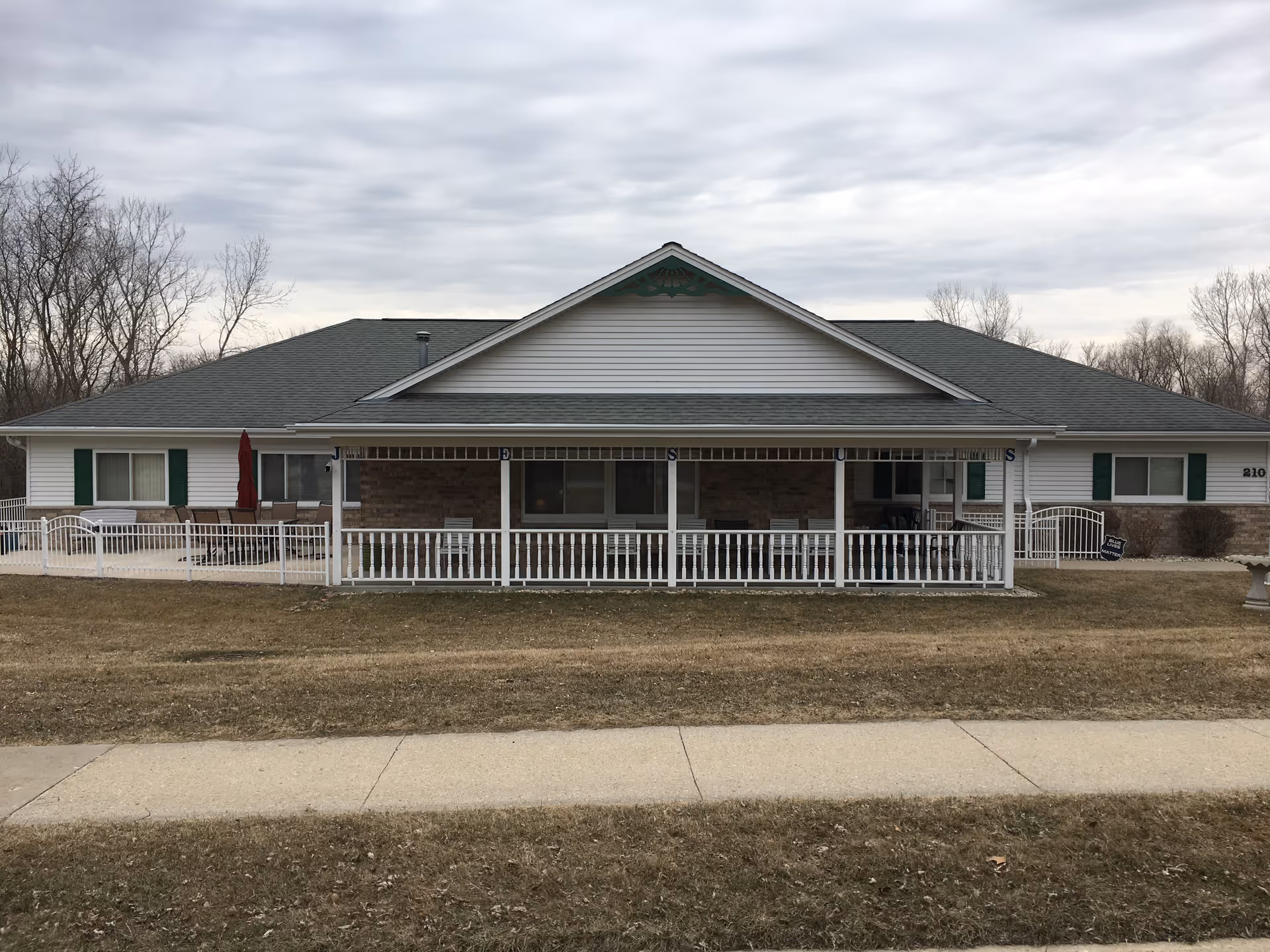 Front exterior view of a single-story senior living facility building with a covered porch, white railing, and green shutters on the windows. The building is surrounded by a lawn and a sidewalk in the foreground, with leafless trees in the background under a cloudy sky.