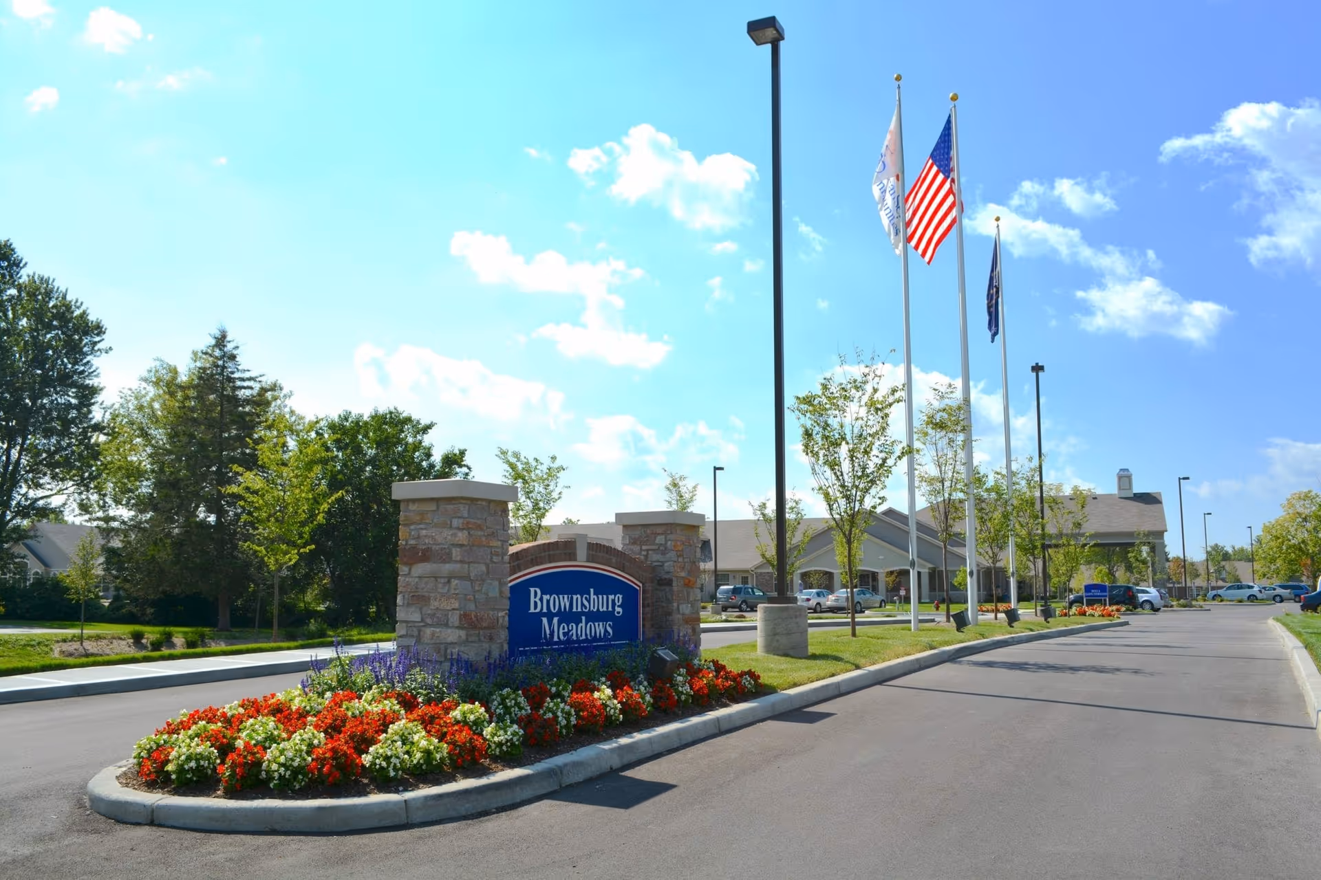 Entrance to Brownsburg Meadows facility with a stone sign surrounded by red, white, and purple flowers. Three flagpoles with flags, a paved driveway, and a building with a covered entrance are visible under a blue sky with scattered clouds.