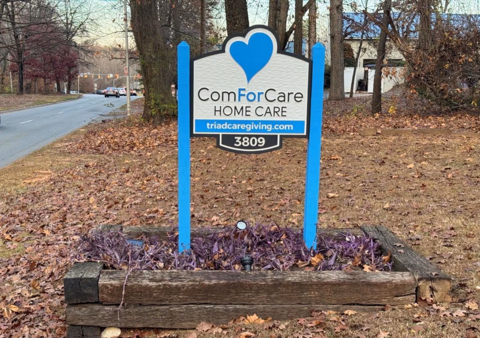 Outdoor ComForCare sign with blue posts in a leaf-covered landscaped bed.