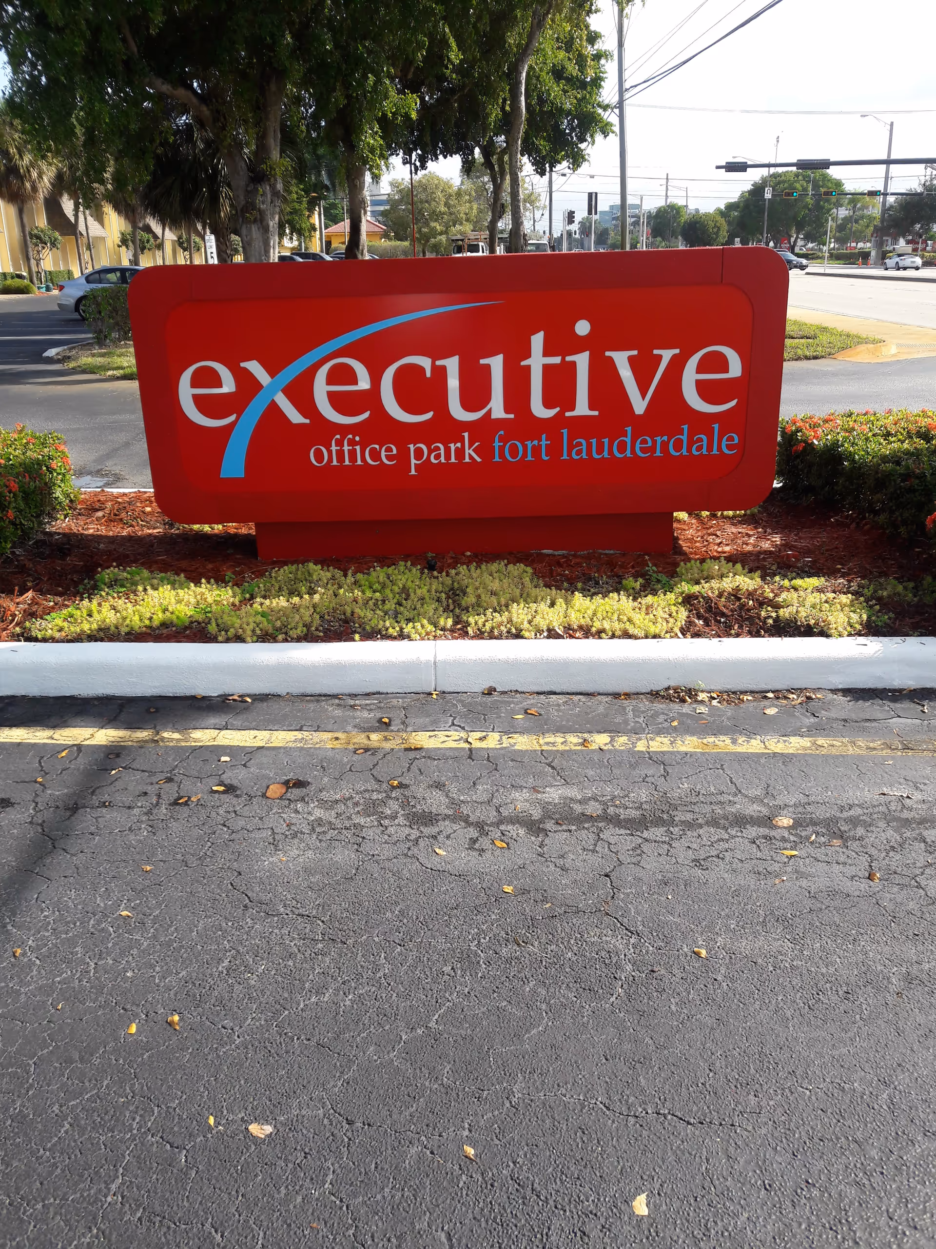 Bright red executive office park sign with landscaped plants beside a street.