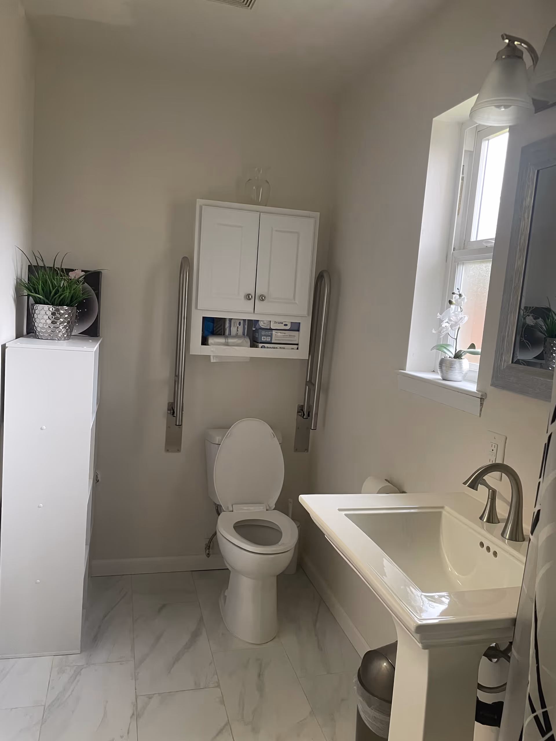 Compact bathroom with toilet, sink, cabinet, grab bars, and a window.