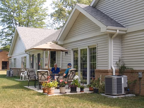 Two people sit at a patio table under an umbrella outside a single-story assisted living building with potted plants and sliding glass doors.