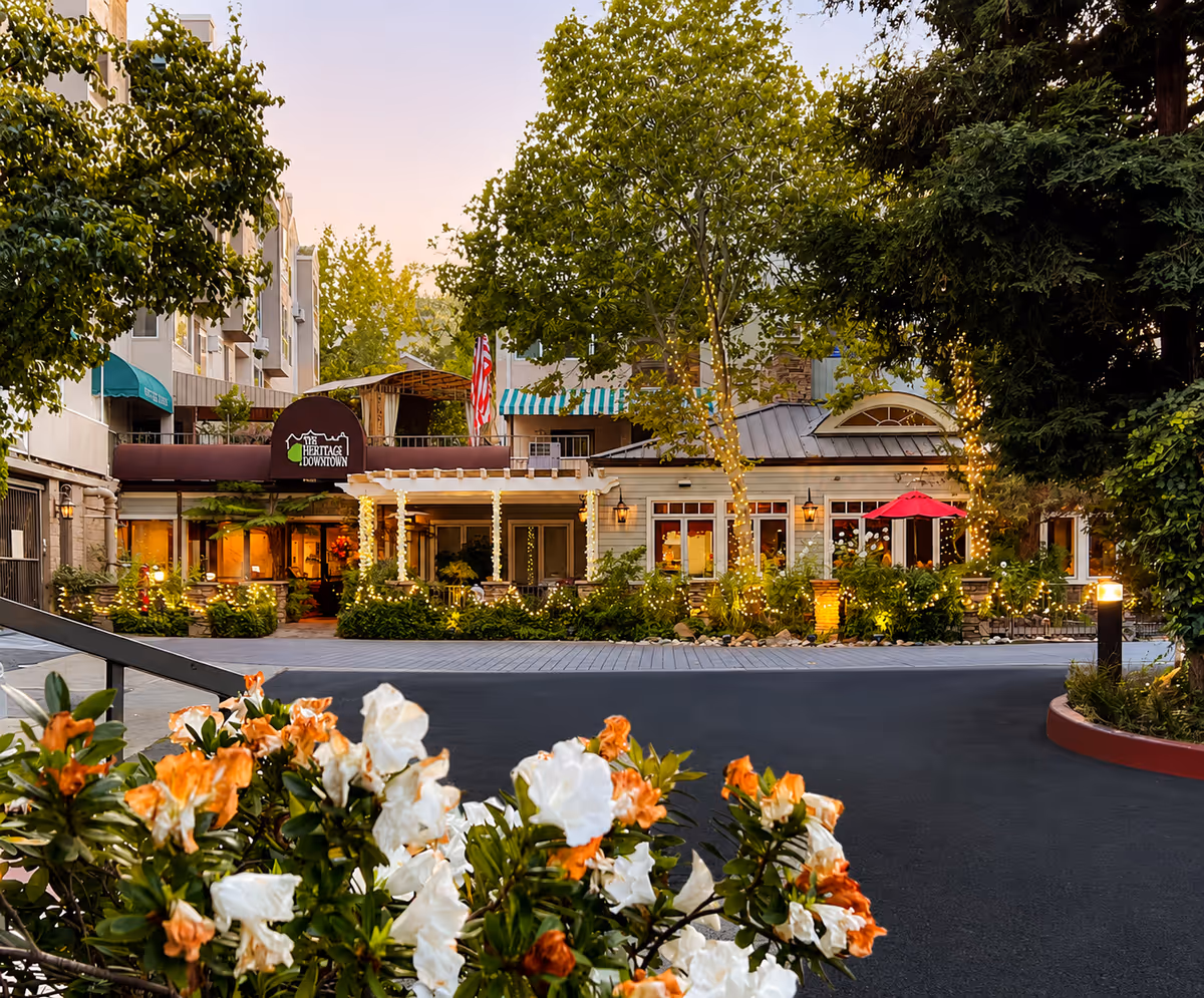 Front exterior of Heritage Downtown senior living with lights, trees, and blooming flowers.