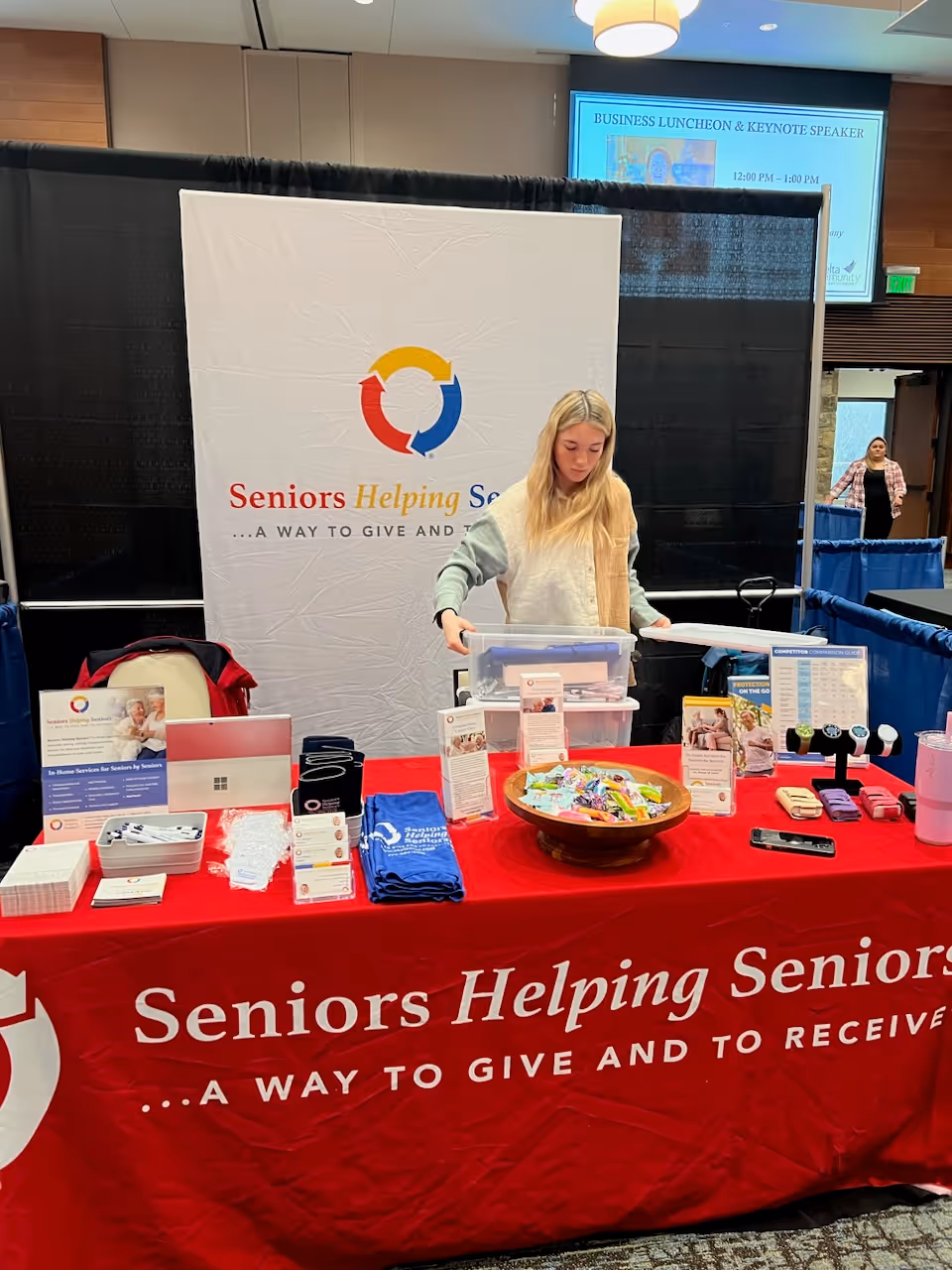 A young woman stands behind a 'Seniors Helping Seniors' booth at an indoor conference.