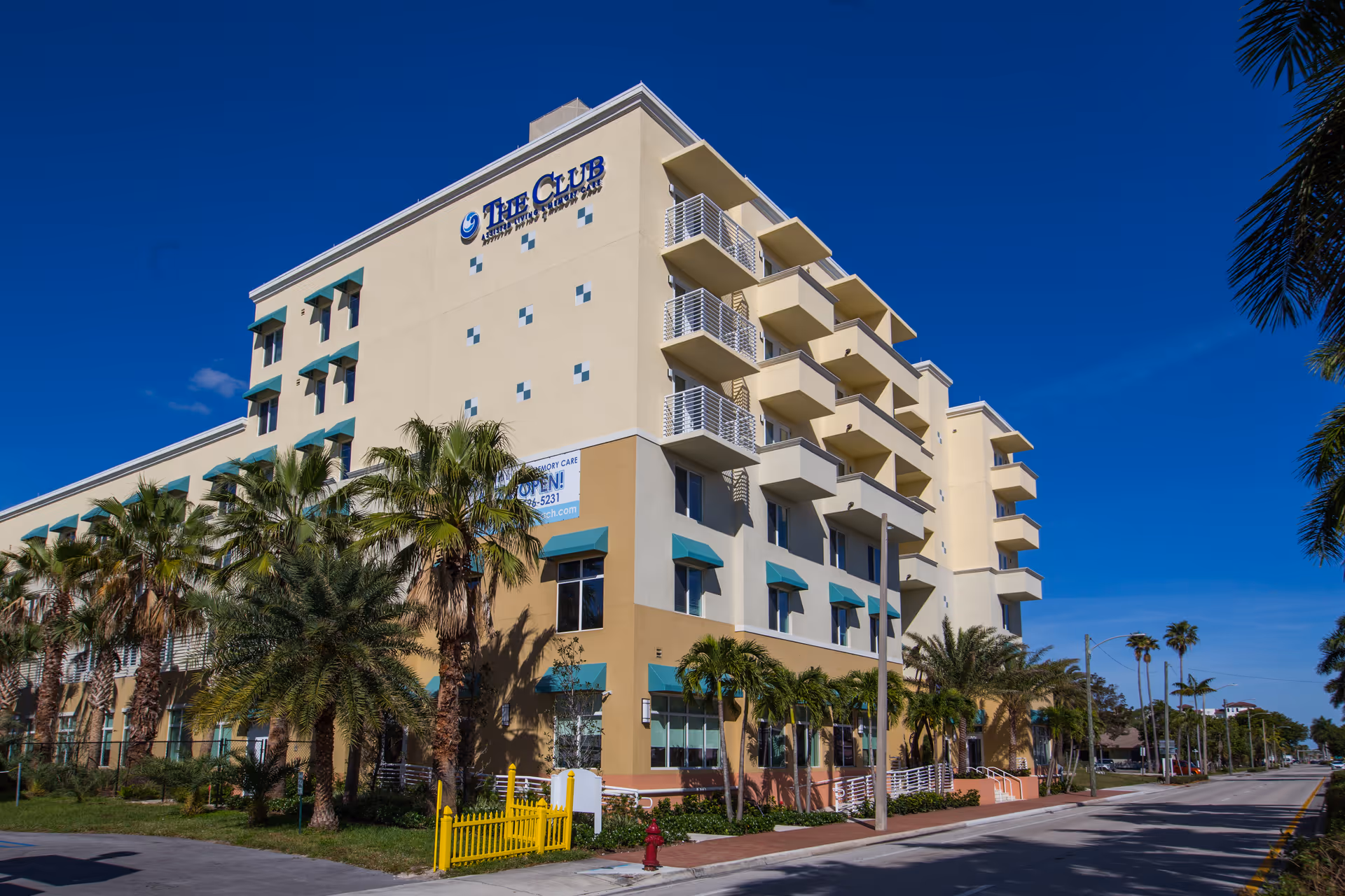 Exterior view of The Club at Boynton Beach, a multi-story senior living facility with balconies, palm trees, and a clear blue sky.