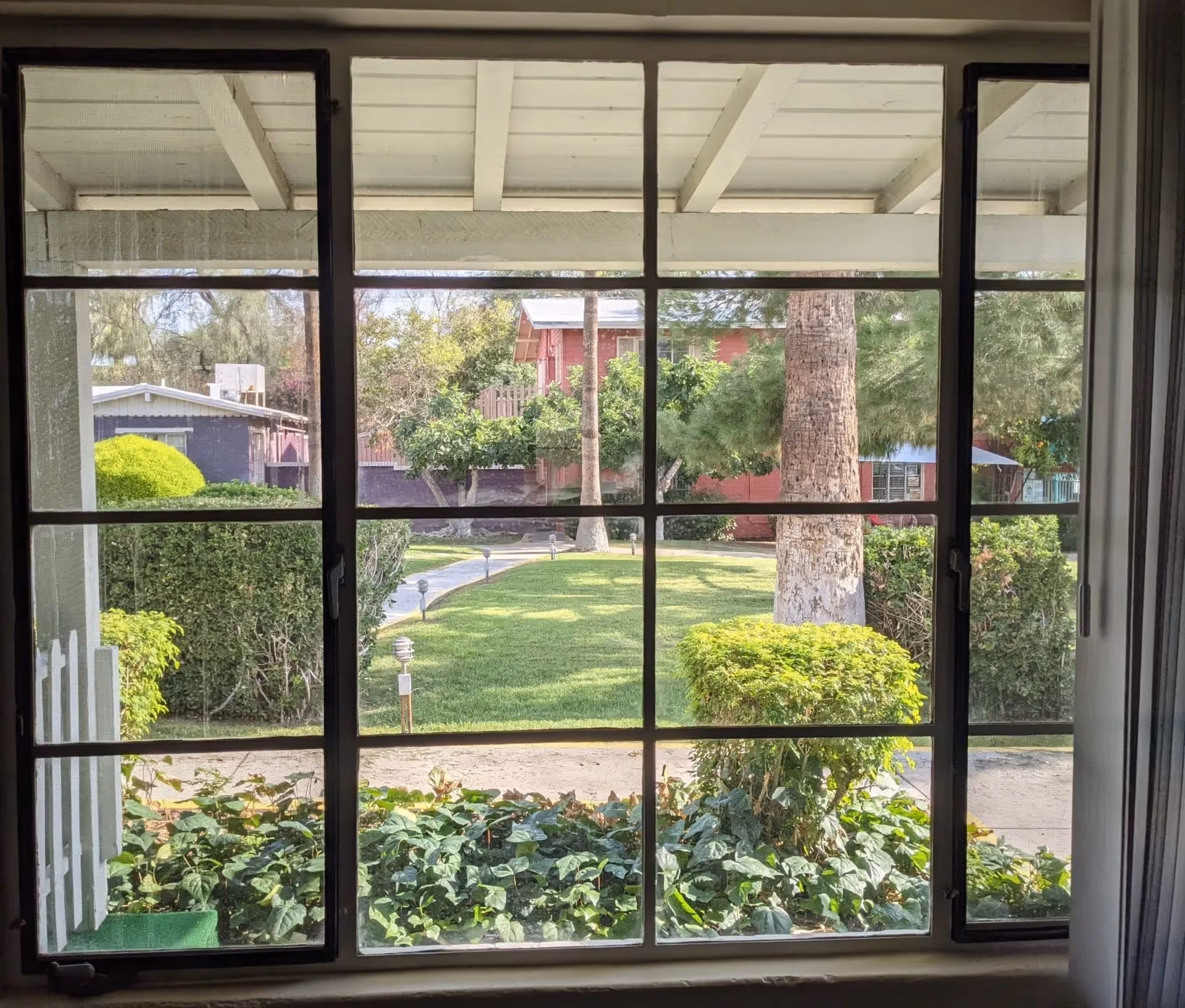 View through a window with black grid frames showing a well-maintained garden outside. The garden has green grass, trimmed bushes, trees, and a paved walkway with small garden lights. A red building and other structures are visible in the background under a white roof overhang.