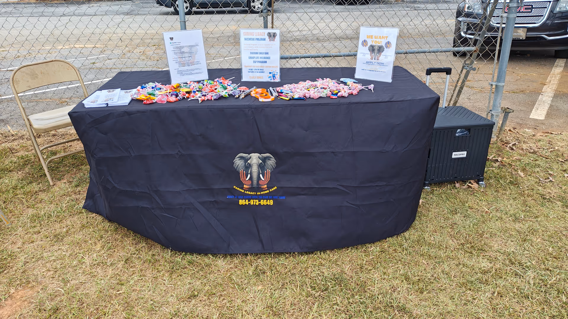 Outdoor event table covered with Caring Legacy LLC branding, candy and flyers, on grass beside chairs.