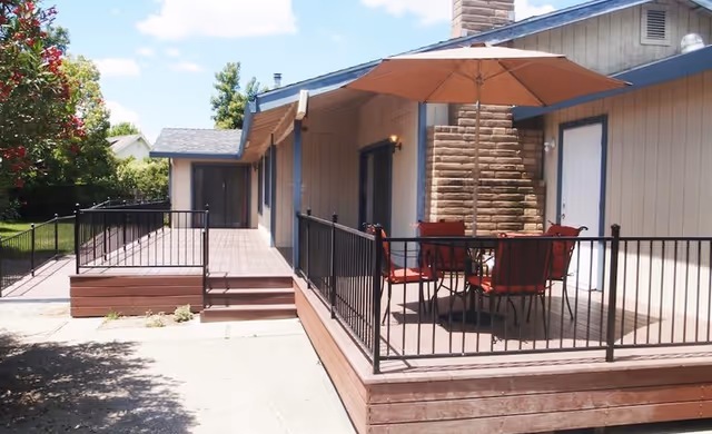 Outdoor patio area with a wooden deck, black metal railing, a table with four red cushioned chairs, and a large beige umbrella. The patio is attached to a beige building with a brick chimney and two doors. Trees and a clear blue sky are visible in the background.