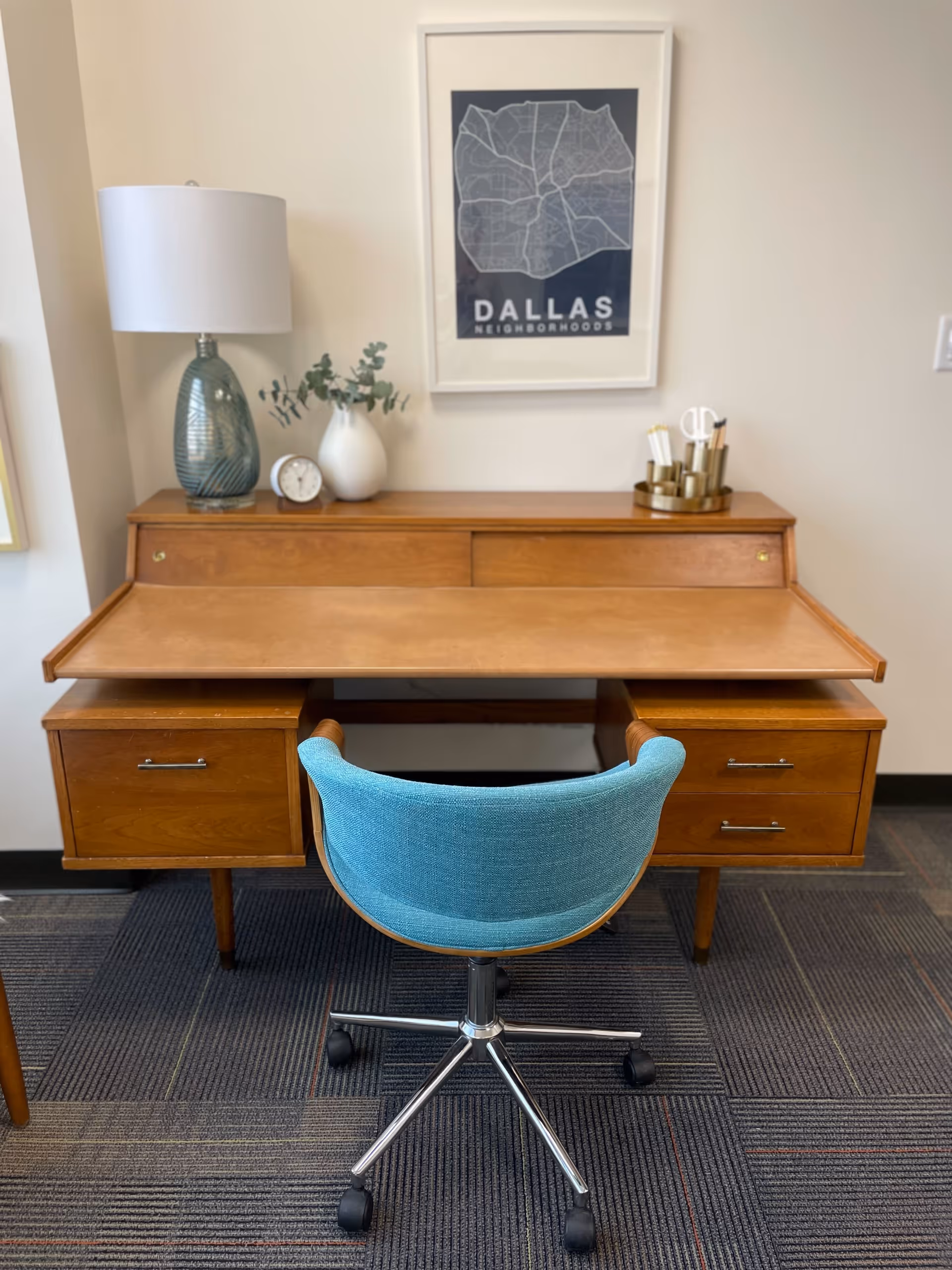 Wooden desk with blue chair, lamp, and framed map poster in a bright office.
