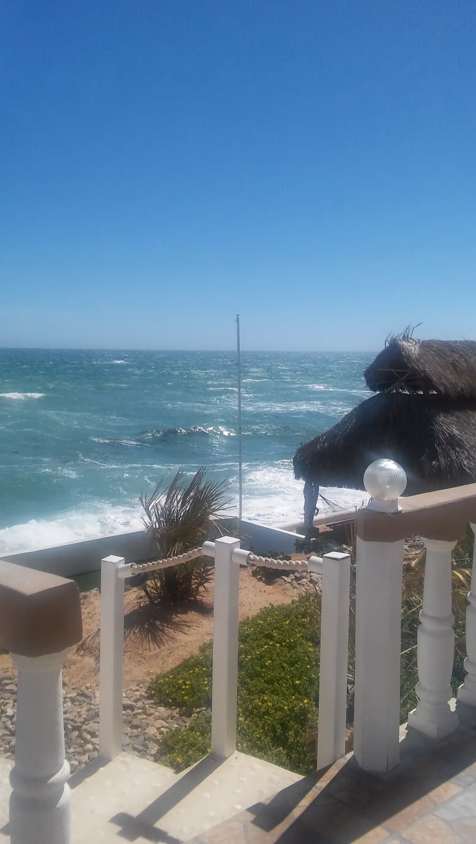 Ocean view from a seaside outdoor patio with white railing and thatched hut.