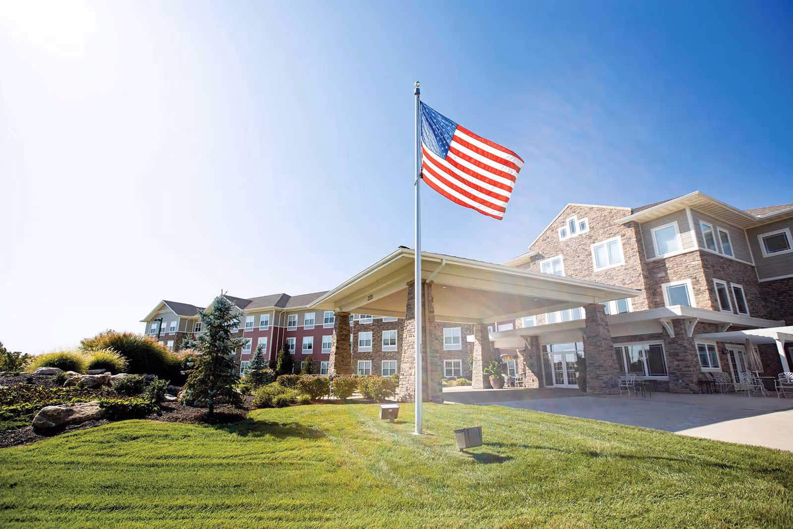 Exterior view of Cedar Creek Of Shiloh facility with a large American flag on a flagpole in front. The building is multi-story with stone and siding exterior, surrounded by well-maintained landscaping and green grass under a clear blue sky.