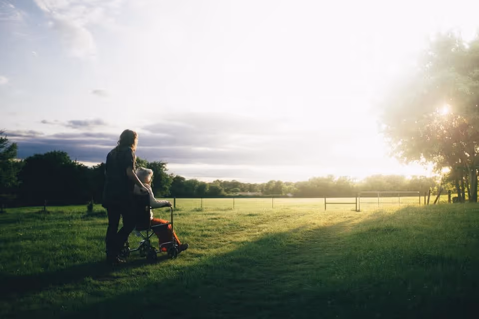 Caregiver pushes an elderly resident in a wheelchair across a sunlit grassy outdoor field.