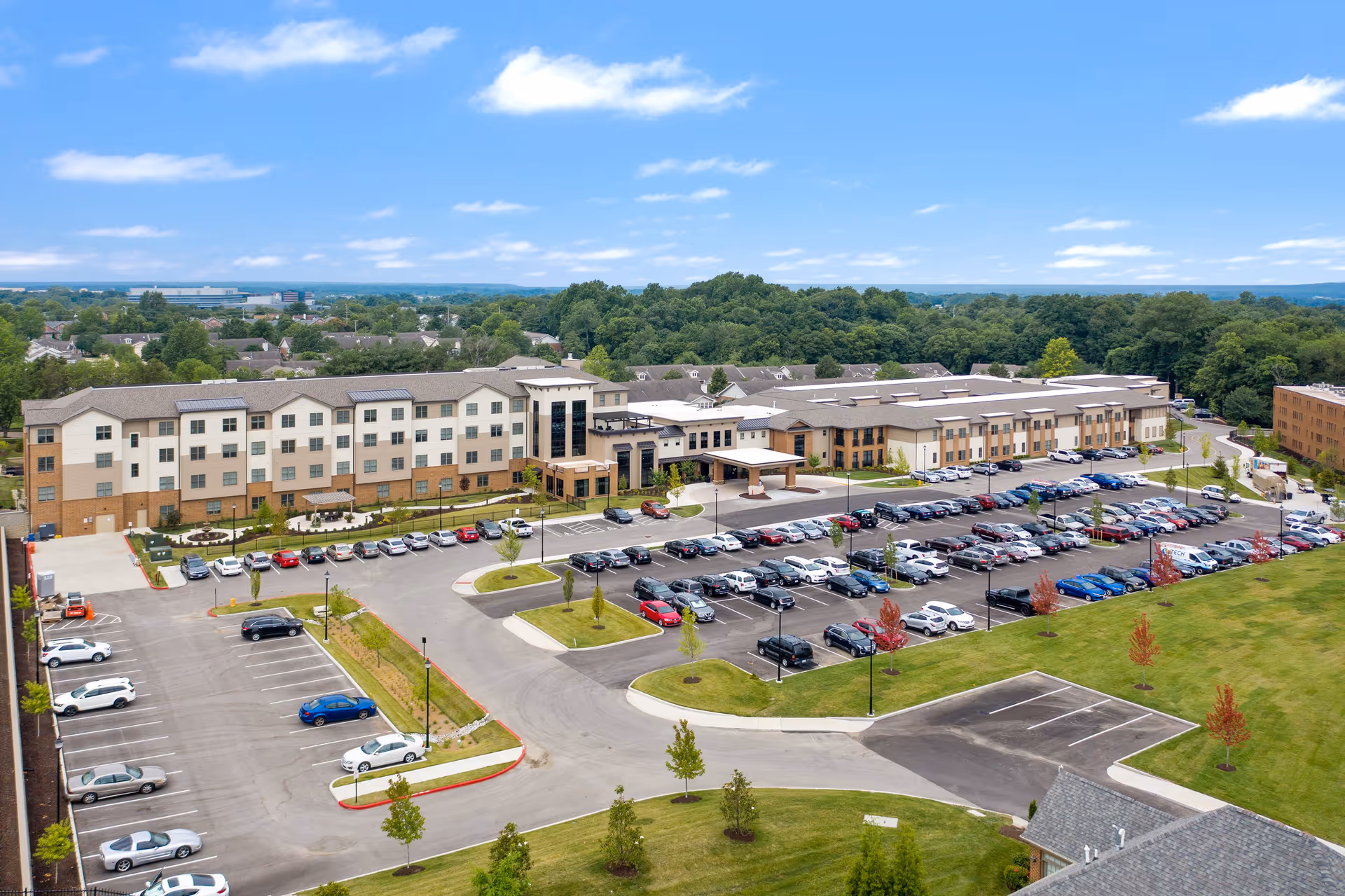 Aerial view of Friendship Village Chesterfield, showing a large multi-story senior living facility with a spacious parking lot filled with cars, surrounded by green lawns and trees under a partly cloudy blue sky.