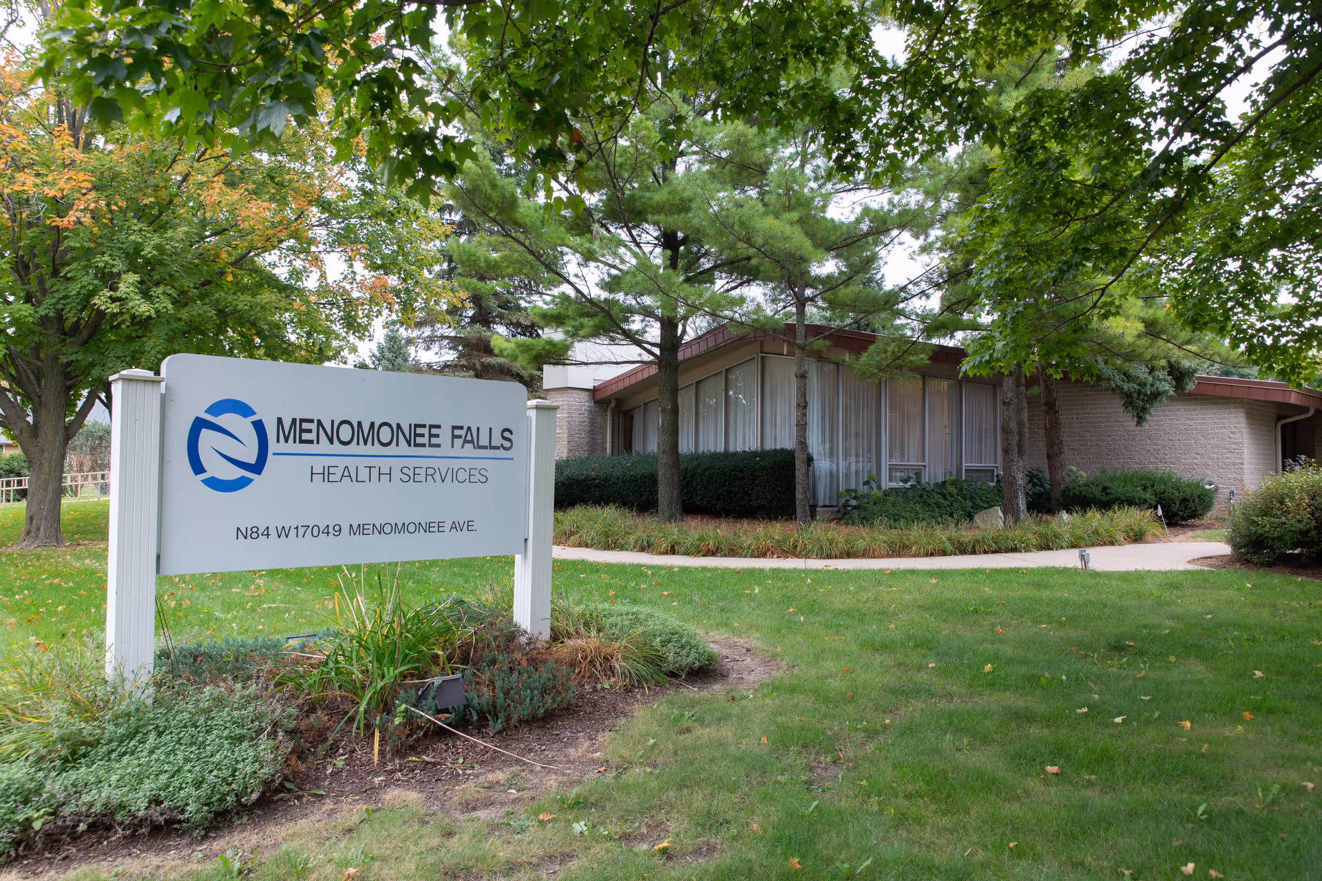 A lawn and trees in front of a single-story building with a sign reading 'Menomonee Falls Health Services'.
