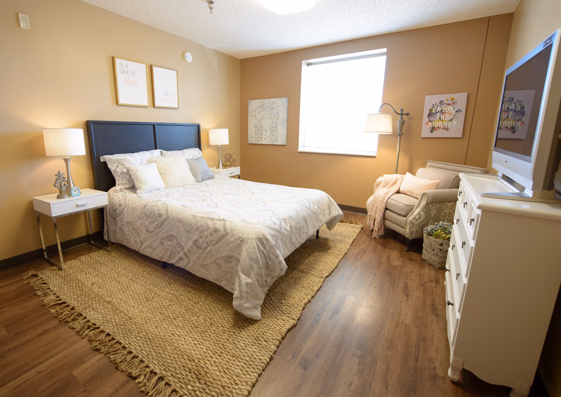 A cozy bedroom with a large bed covered in white patterned bedding, two bedside tables with lamps, a beige armchair with a pink throw blanket, a white dresser with a TV on top, and wall art including framed quotes and decorative pieces. The room has warm beige walls and wooden flooring with a woven rug under the bed.