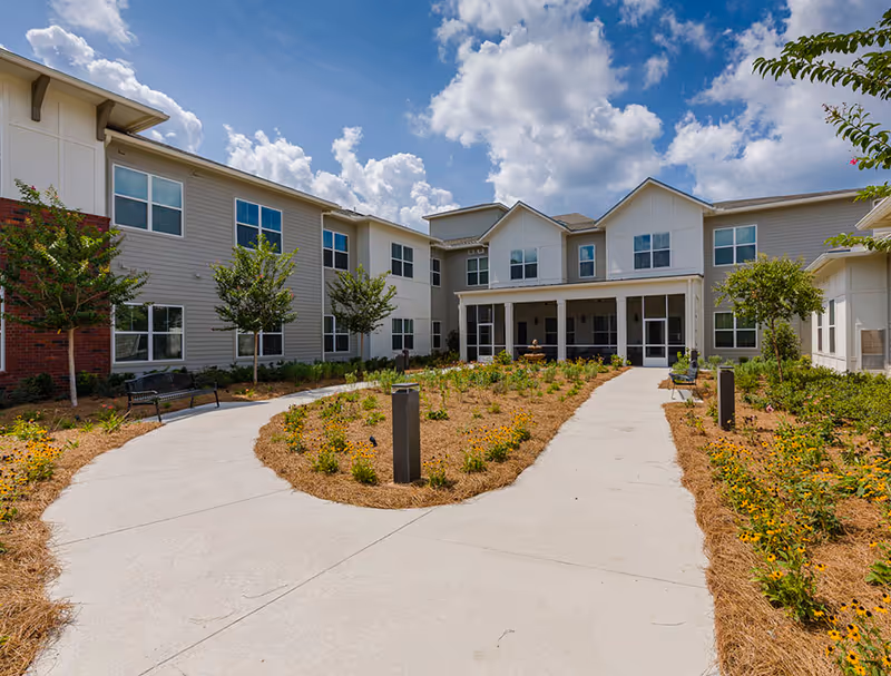 Exterior view of a two-story senior living facility building with beige siding and white trim. The building surrounds a landscaped courtyard with young trees, shrubs, and a concrete walkway leading to a covered entrance. The sky is partly cloudy with bright sunlight.