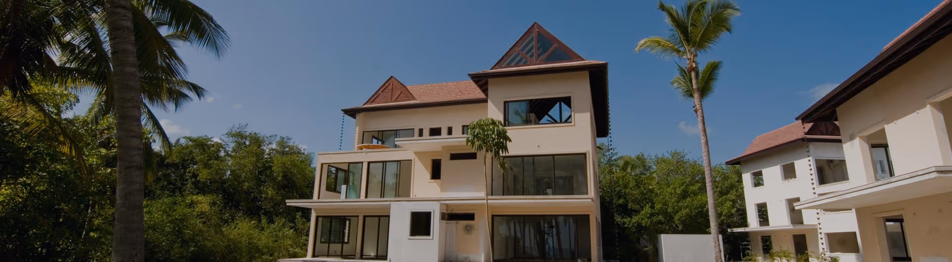 Multi-story beige residential building with balconies and large windows framed by palm trees under a clear blue sky.