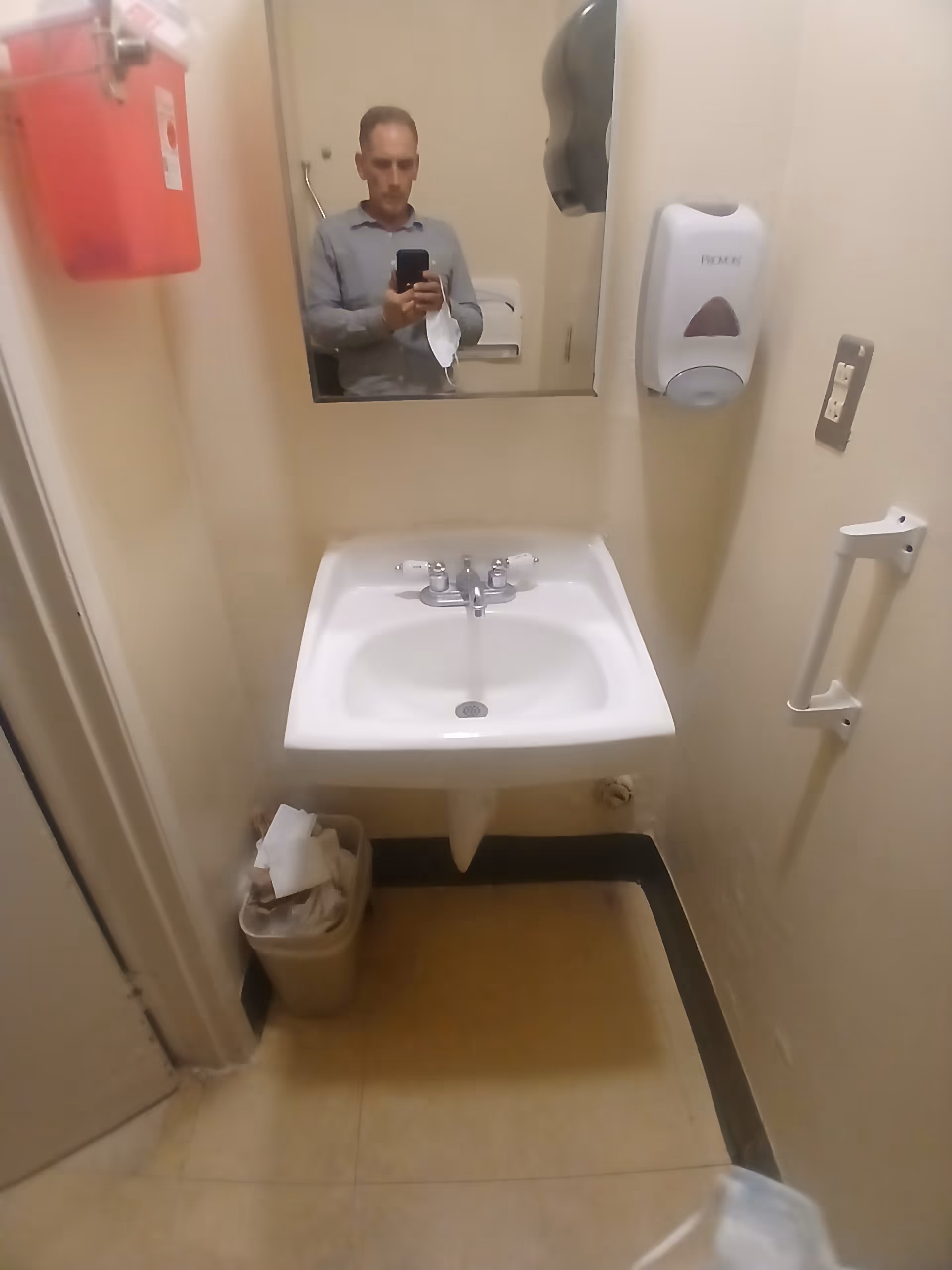 Small bathroom with sink, mirror, soap dispenser, and a man taking a selfie.