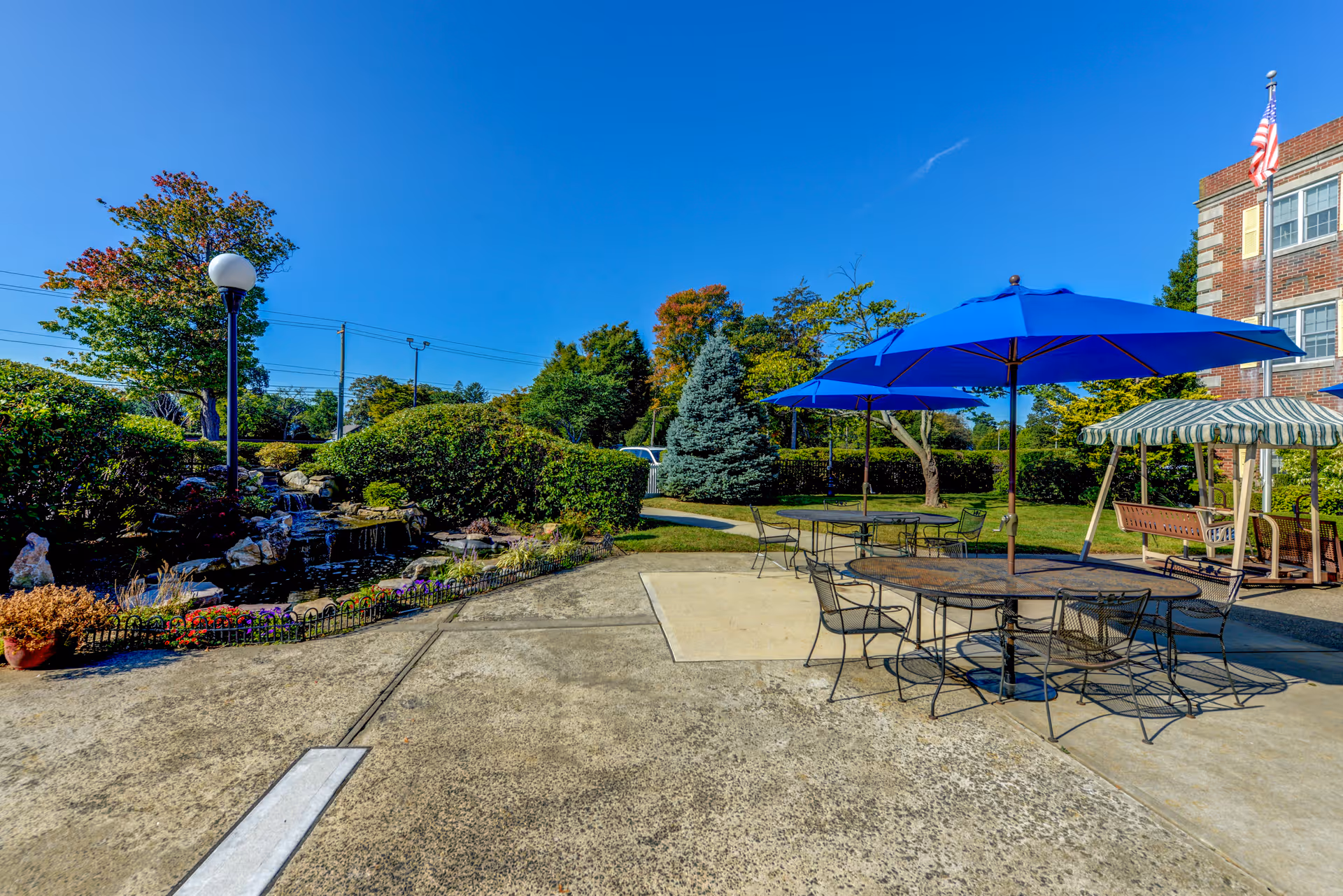 Outdoor patio with metal tables and blue umbrellas beside a landscaped pond and a brick building.
