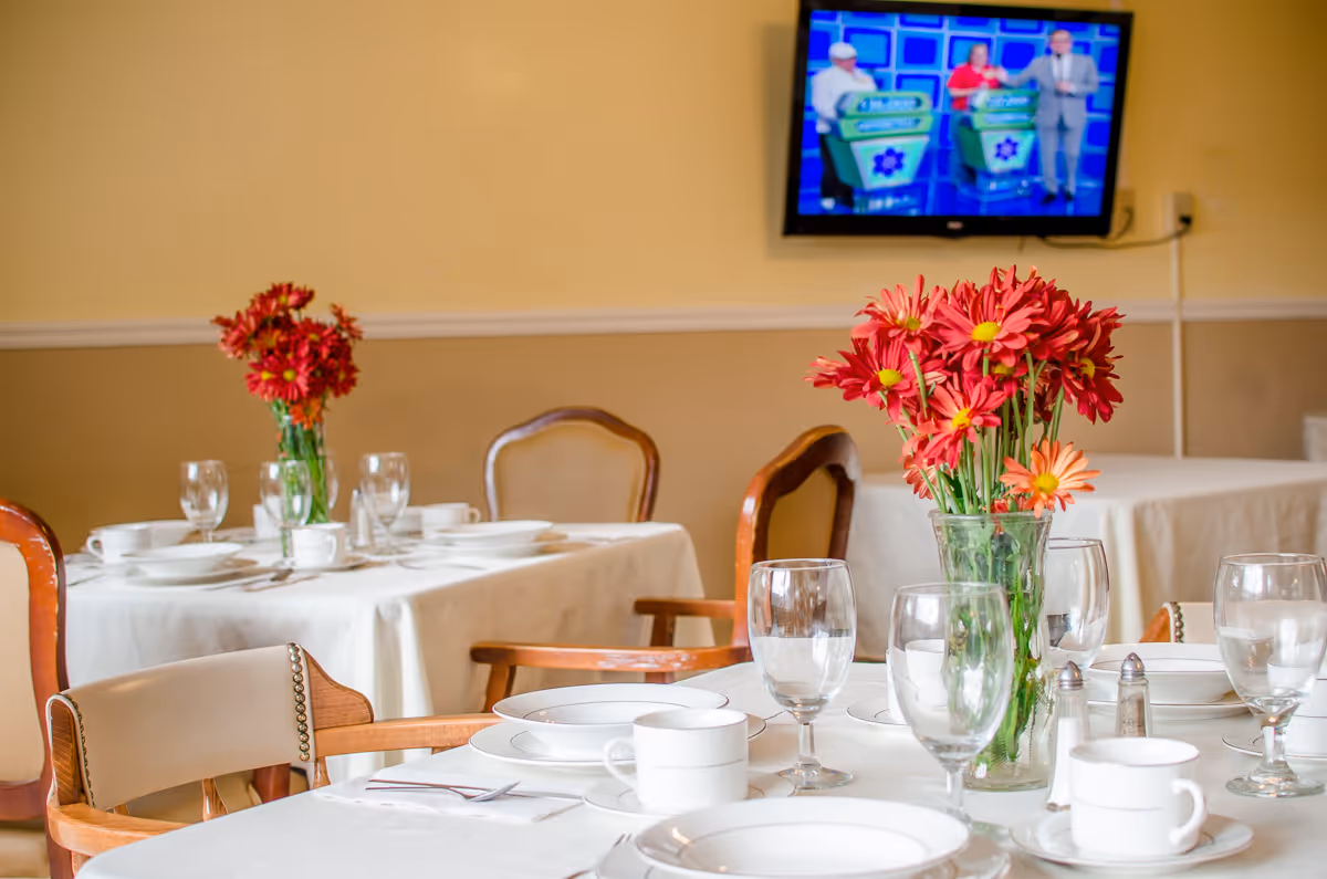 Dining room with white tablecloths, flowers, glassware, and a wall mounted TV.