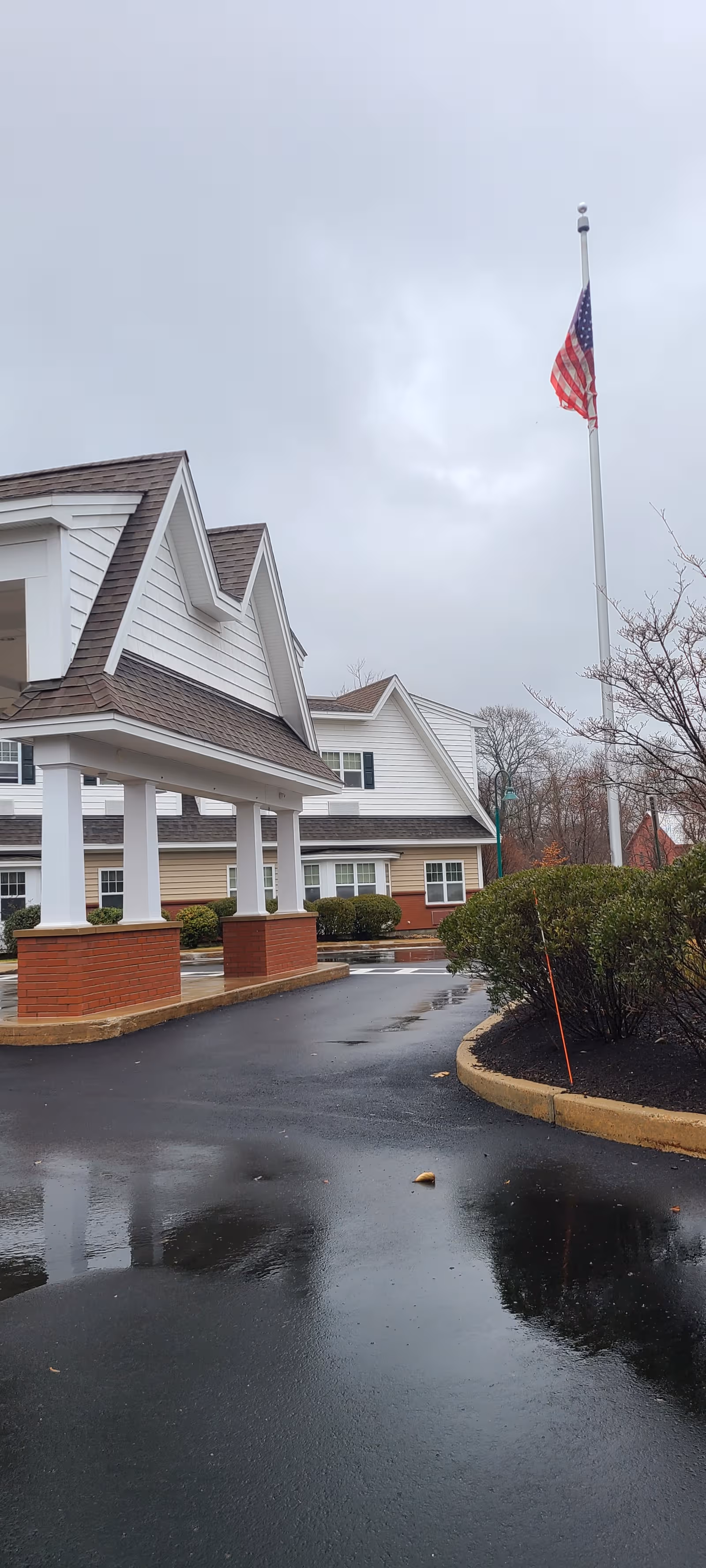 Senior living facility exterior with a porte-cochere, flagpole, and wet pavement.